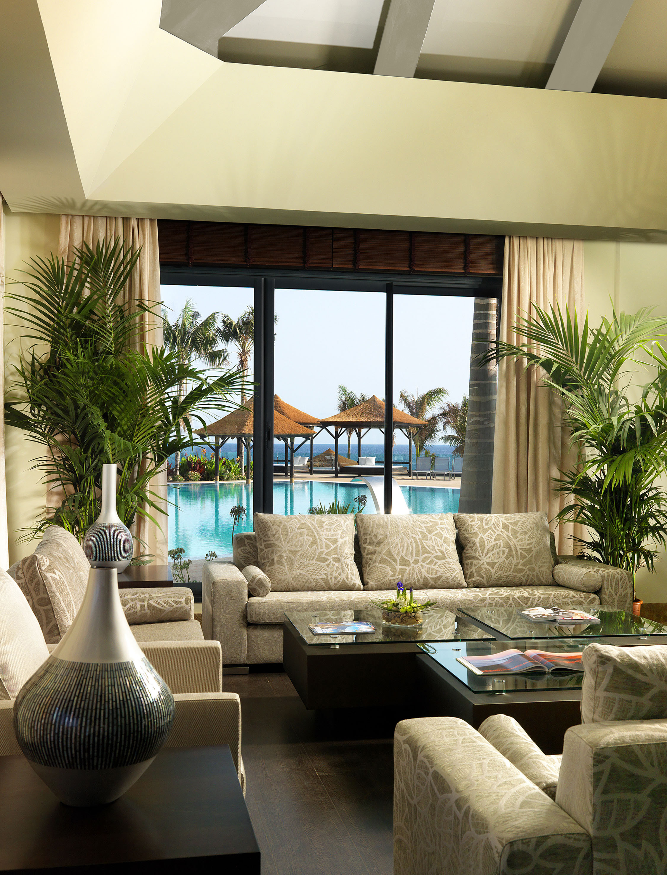 Luxury resort lounge with tropical plants, glass tables, and pool-ocean view.