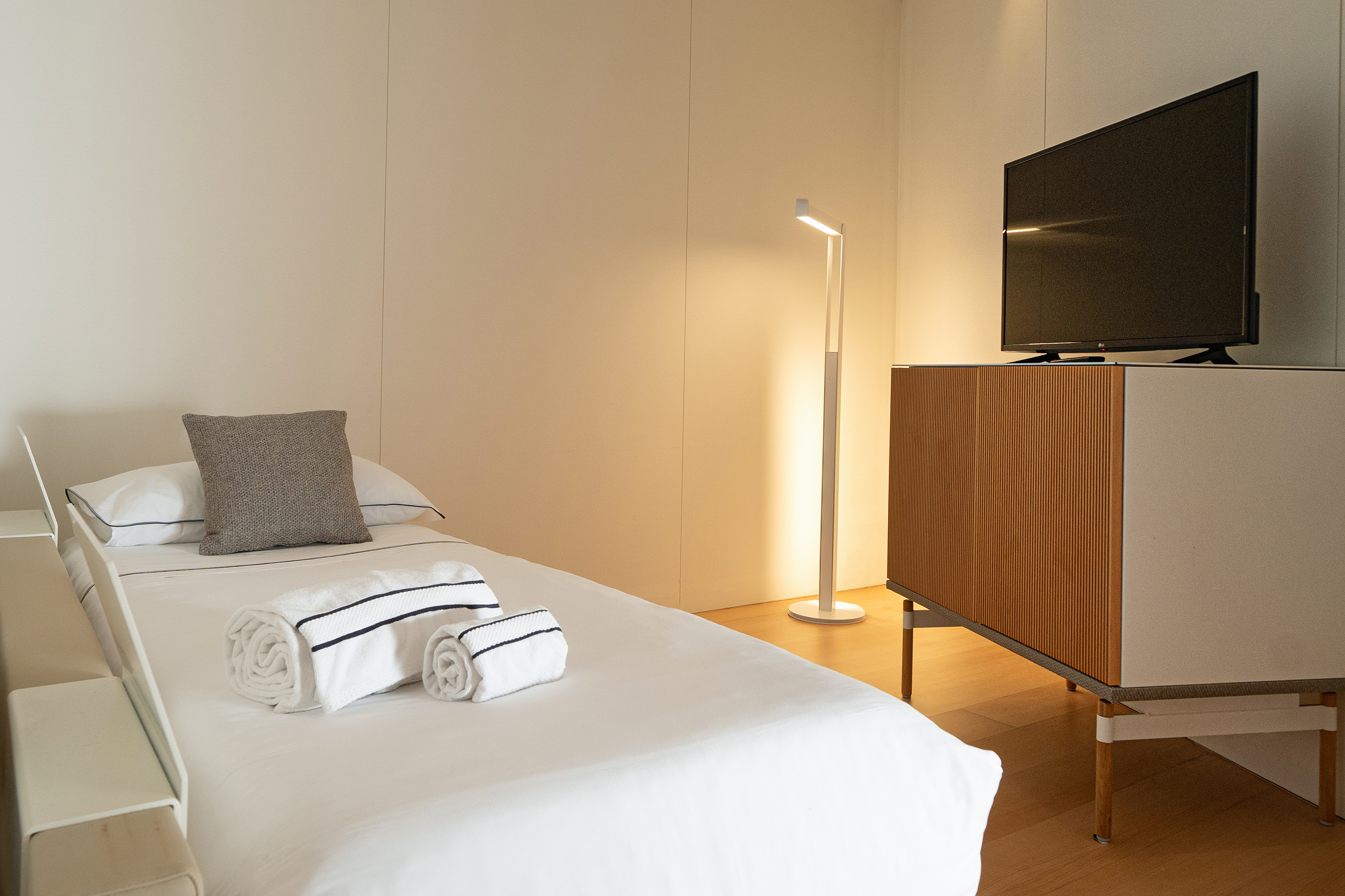 Modern minimalist bedroom with white bed, rolled towels, floor lamp, and TV.