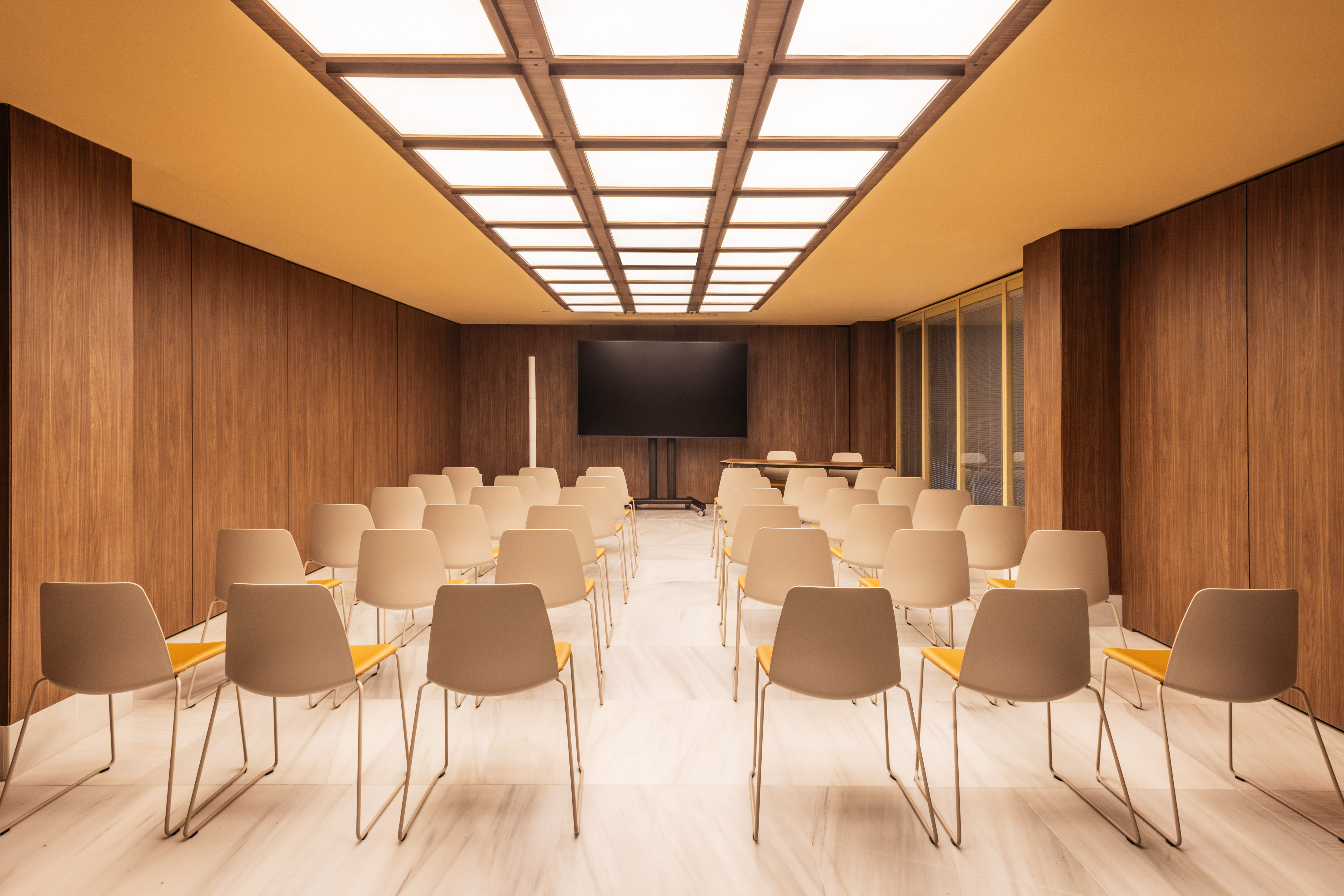 Modern meeting room with beige chairs, large screen, wood walls, ceiling lights.