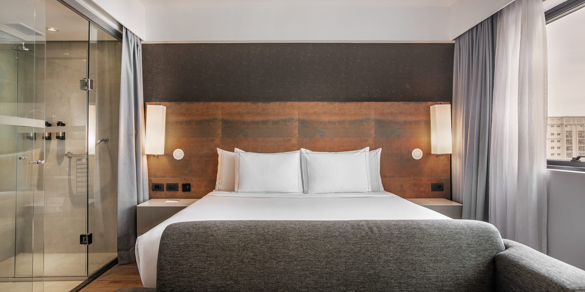 Quarto de hotel moderno com cama de casal, sofá cinza e banheiro com parede de vidro.