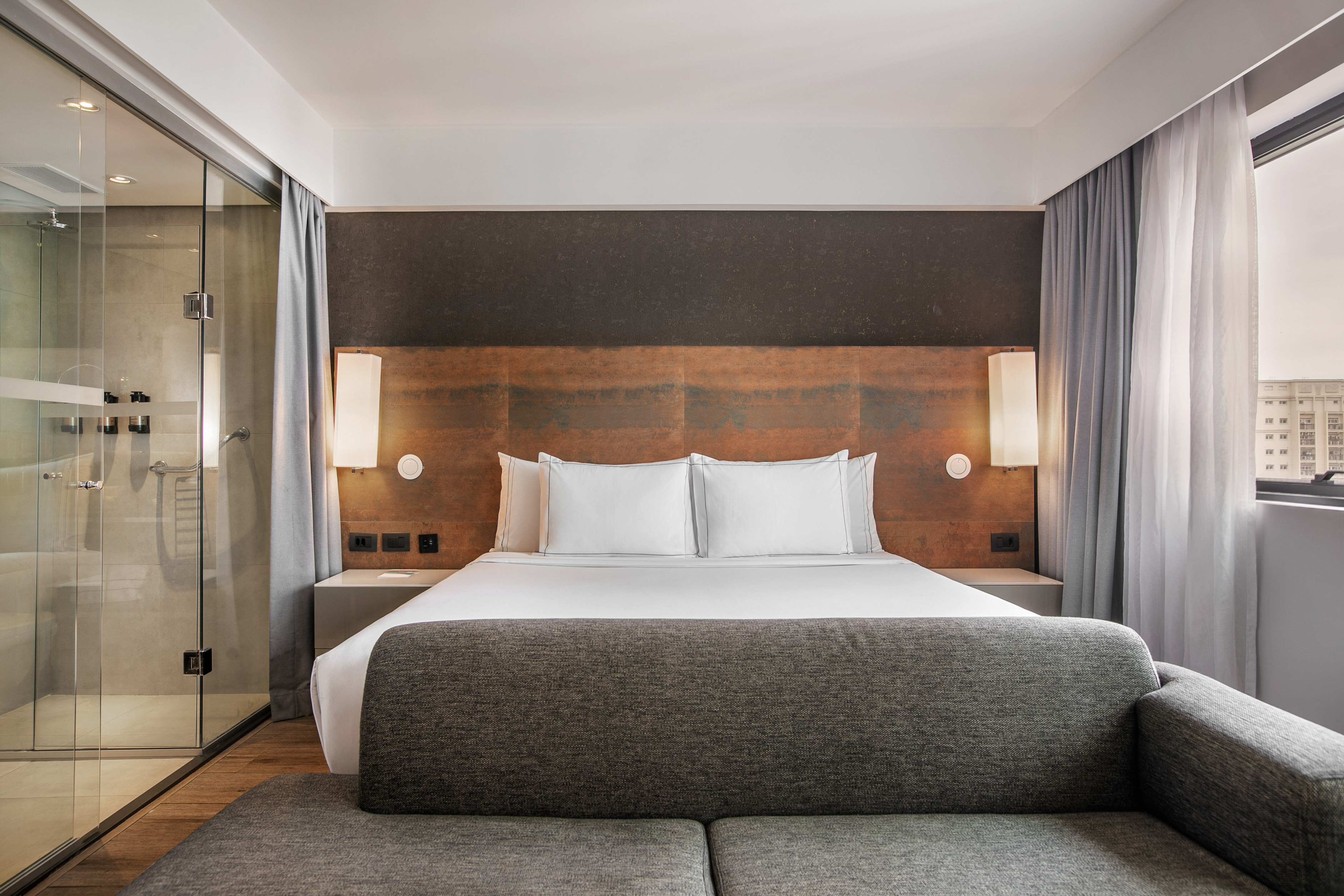 Quarto de hotel moderno com cama grande, cabeceira de madeira, banheiro de vidro e sofá cinza. - Grand Premium - Meliá Ibirapuera, São Paulo
