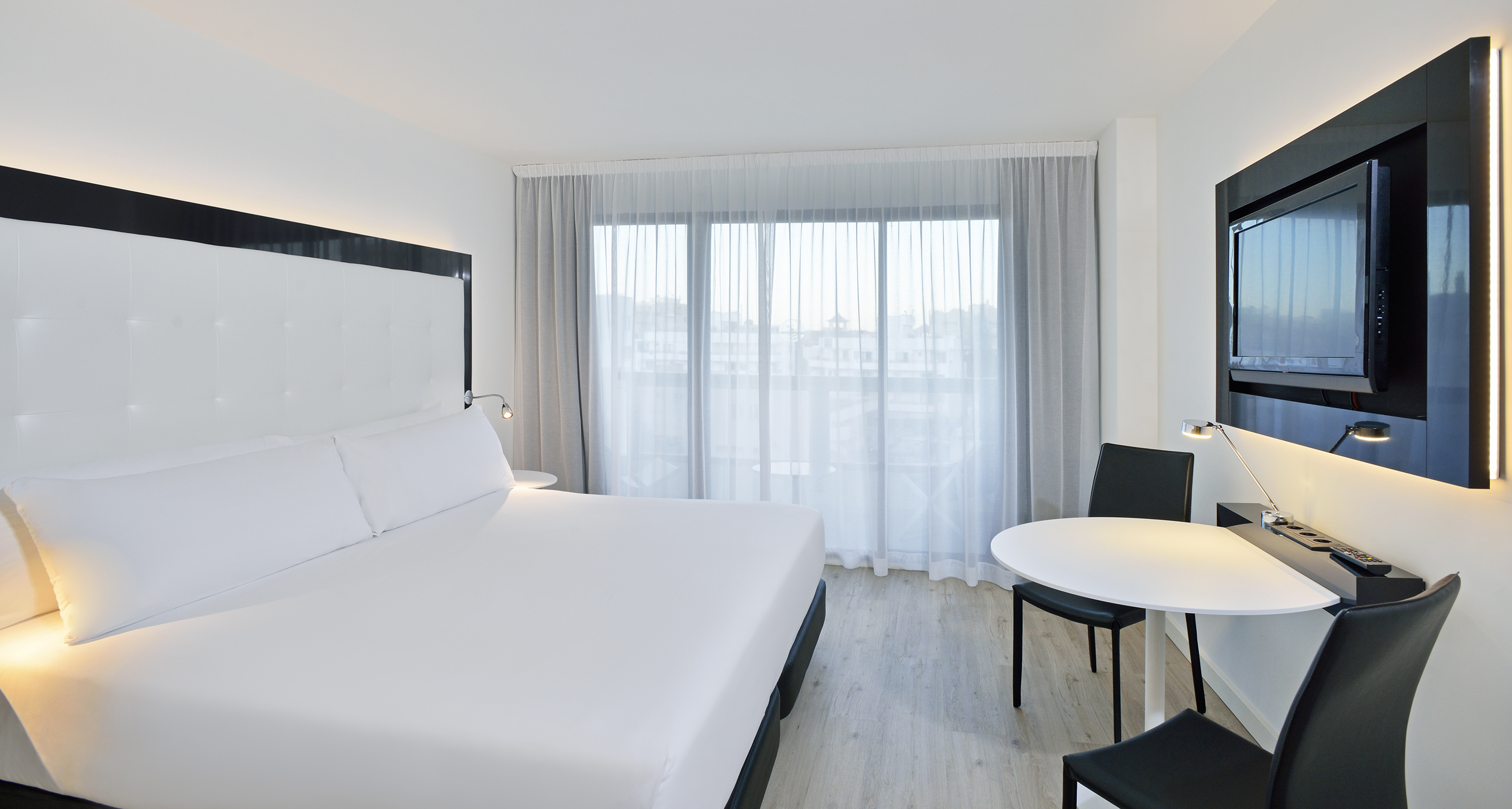 Modern hotel room with white bed, large window, wall-mounted TV, and desk.