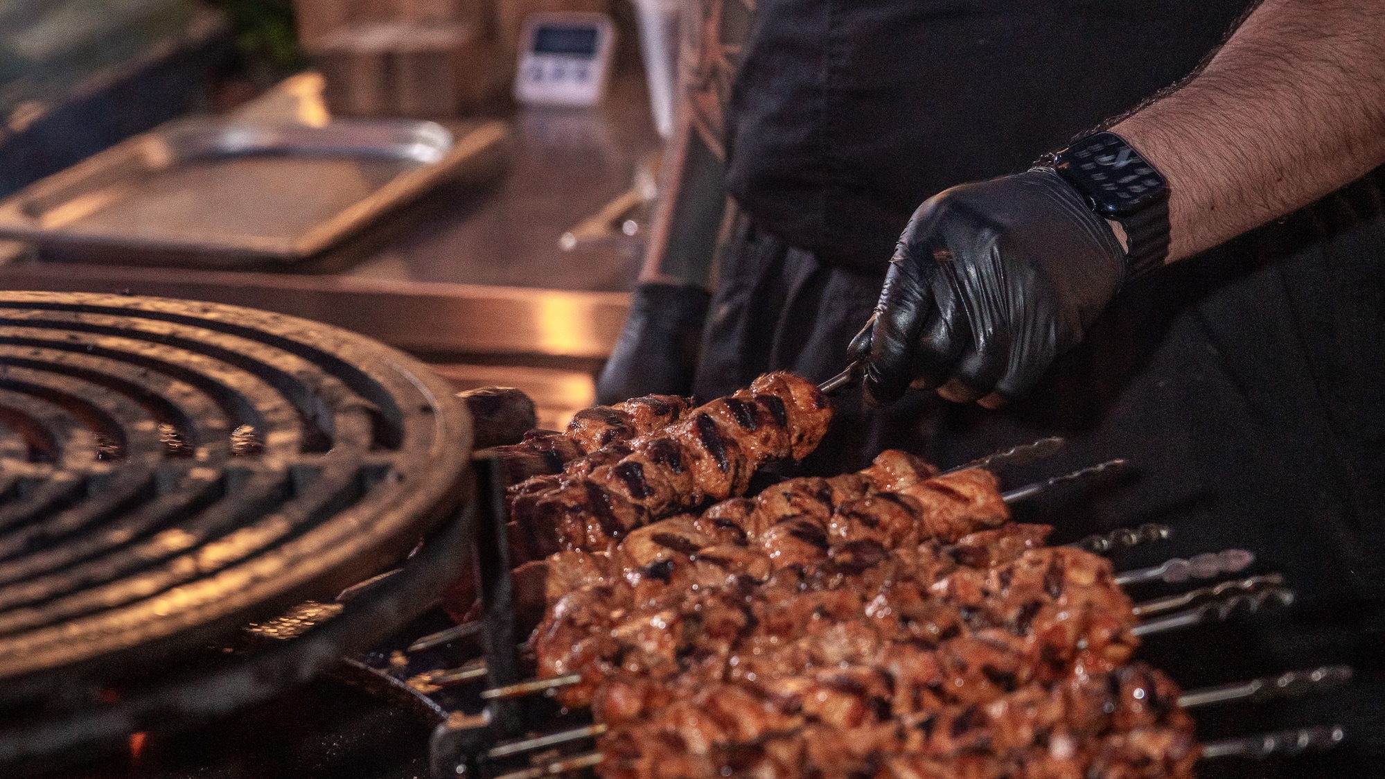 Gloved chef grilling savory meat skewers over hot coals on a BBQ.