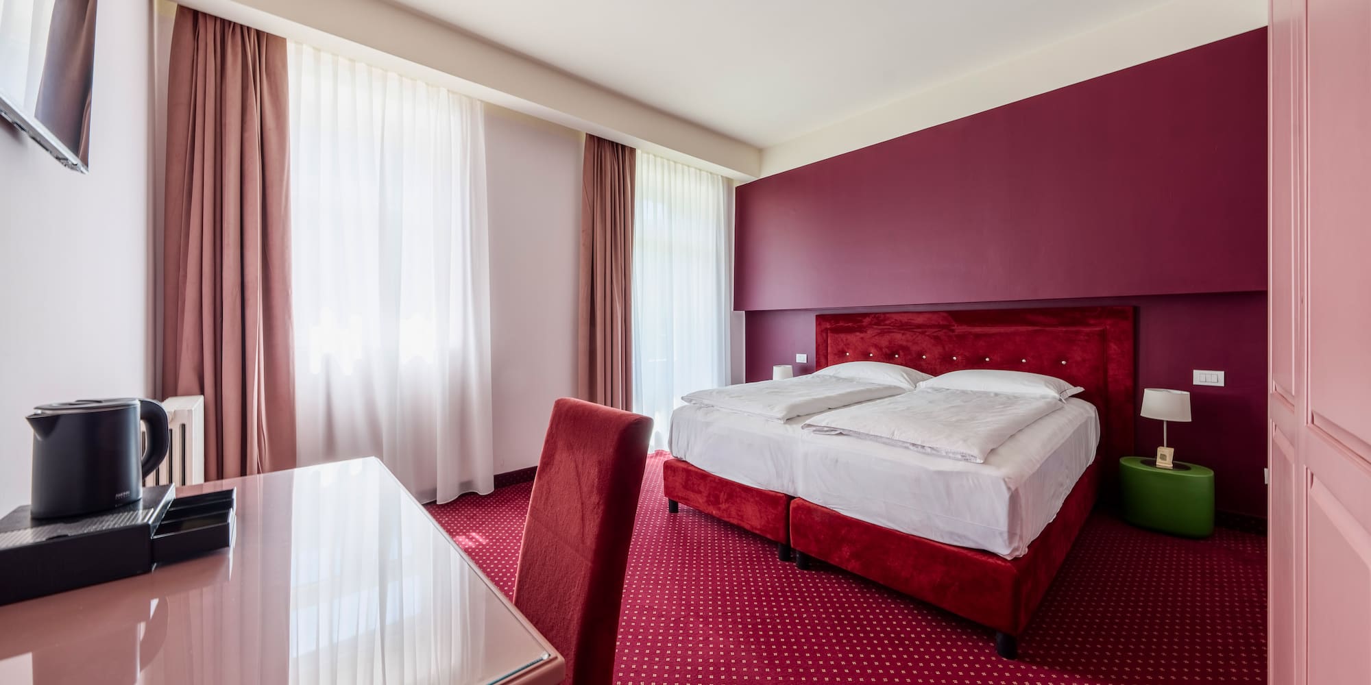 Modern hotel bedroom with red bed, patterned carpet, accent wall, and desk.