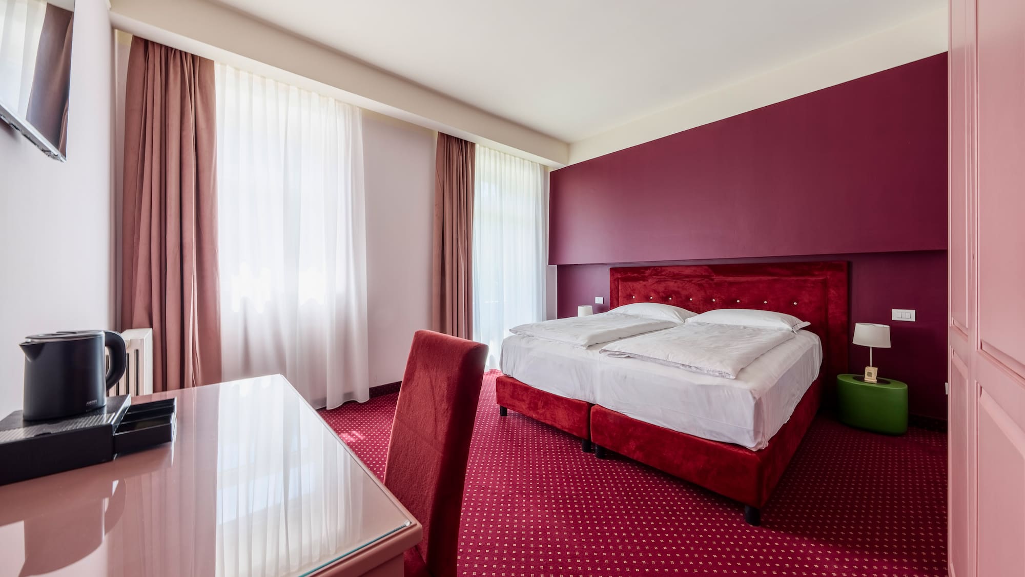 Red hotel room with large bed, patterned carpet, desk, and burgundy wall.