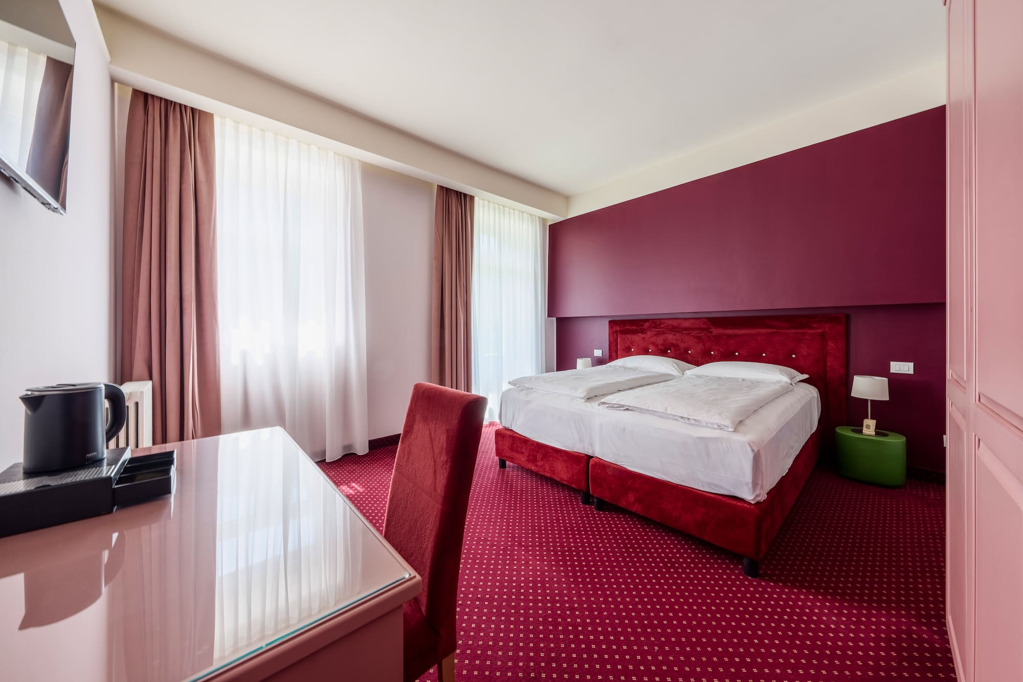 Modern hotel bedroom with red bed, patterned carpet, accent wall, and desk.