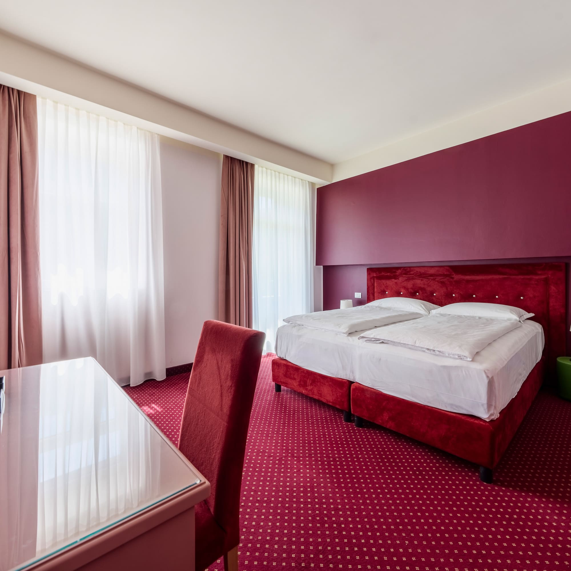 Modern hotel bedroom with red bed, patterned carpet, accent wall, and desk.