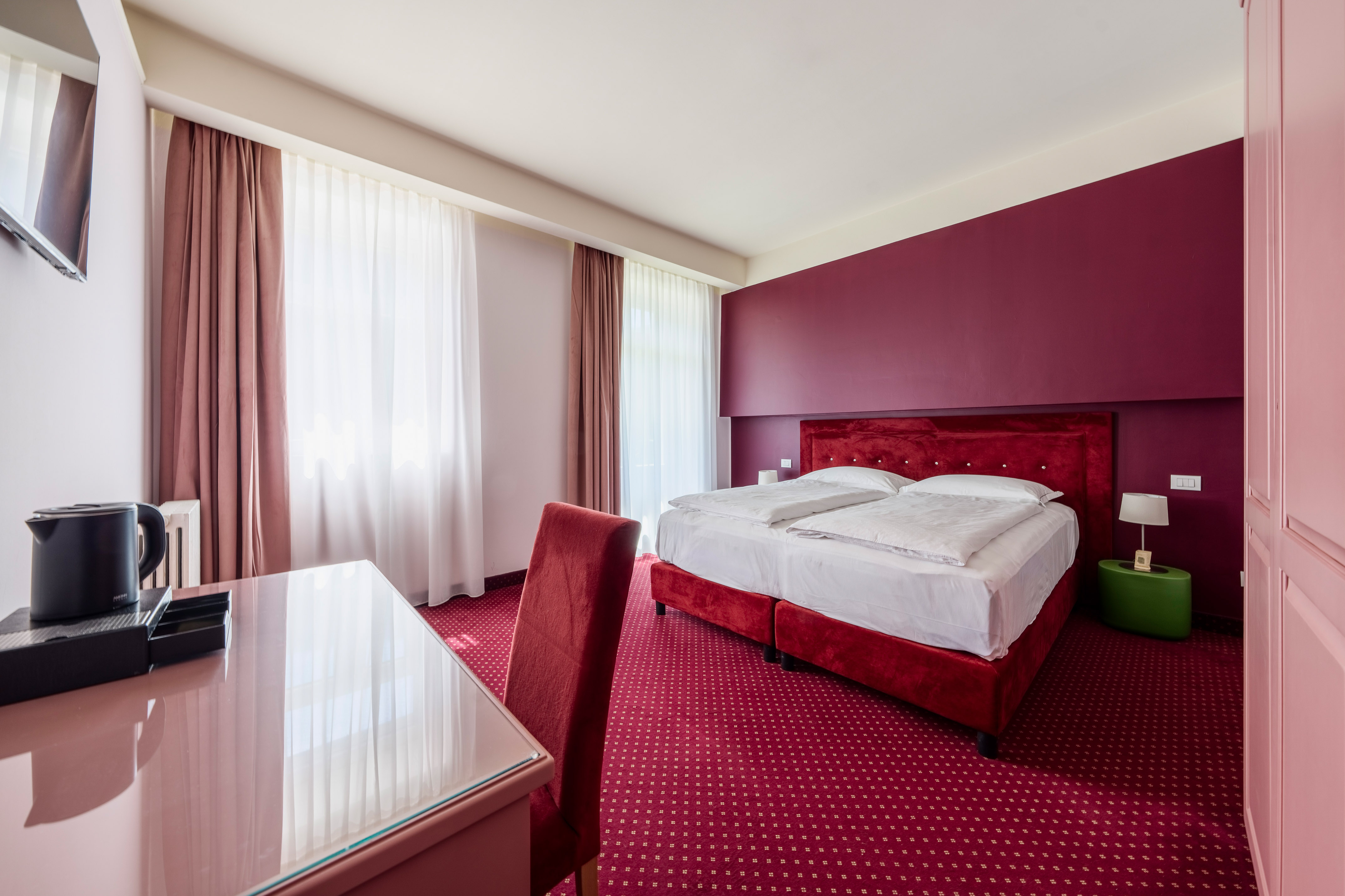 Modern hotel bedroom with red bed, patterned carpet, accent wall, and desk.
