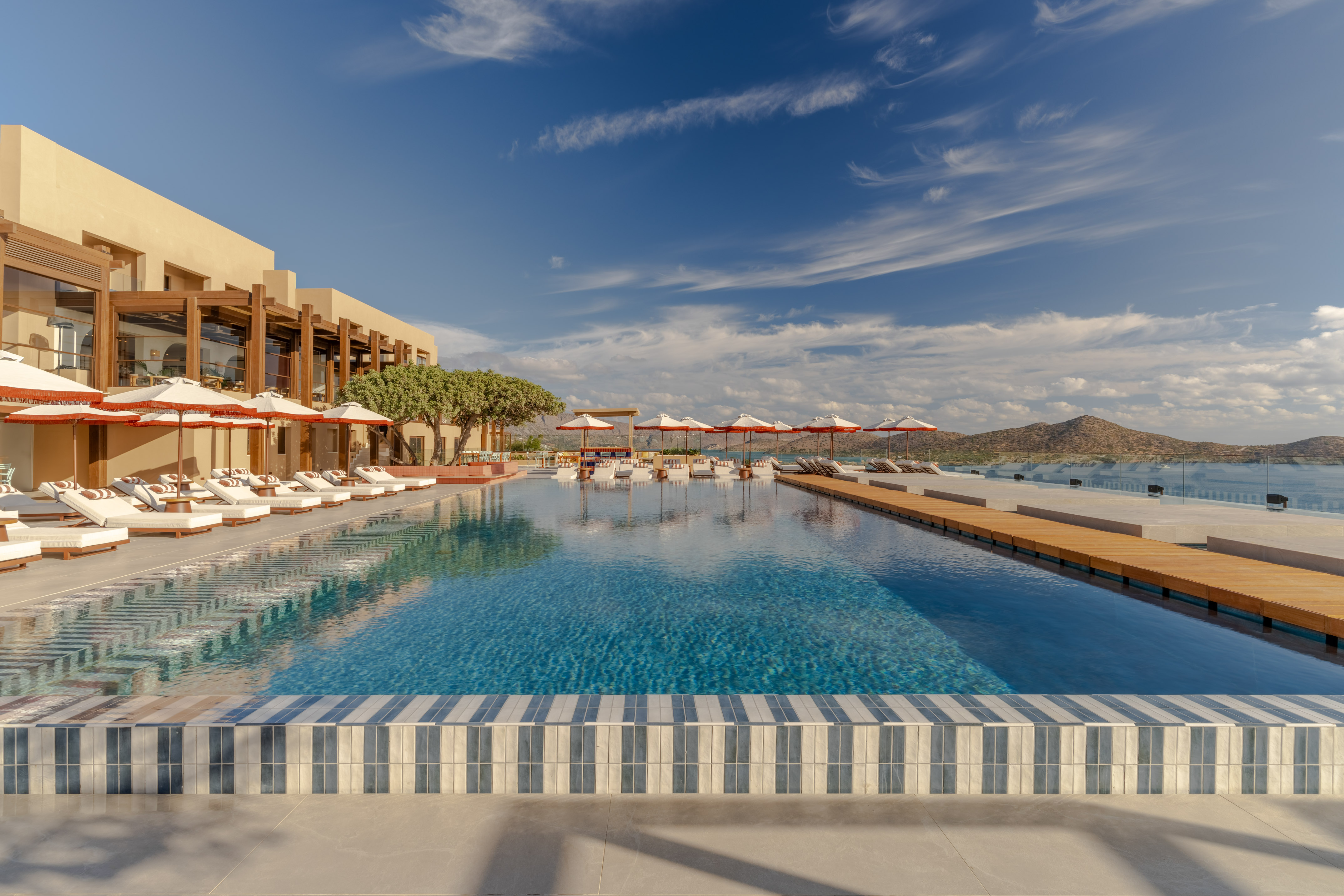 Luxury resort infinity pool with ocean views, sun loungers, and mountains.