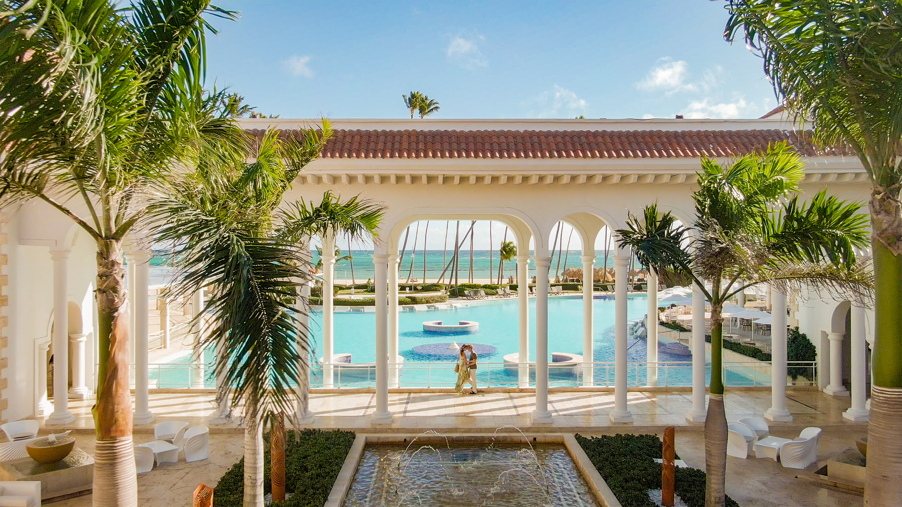 Tropical resort: couple by large pool, ocean, beach, palm trees, and white arches.