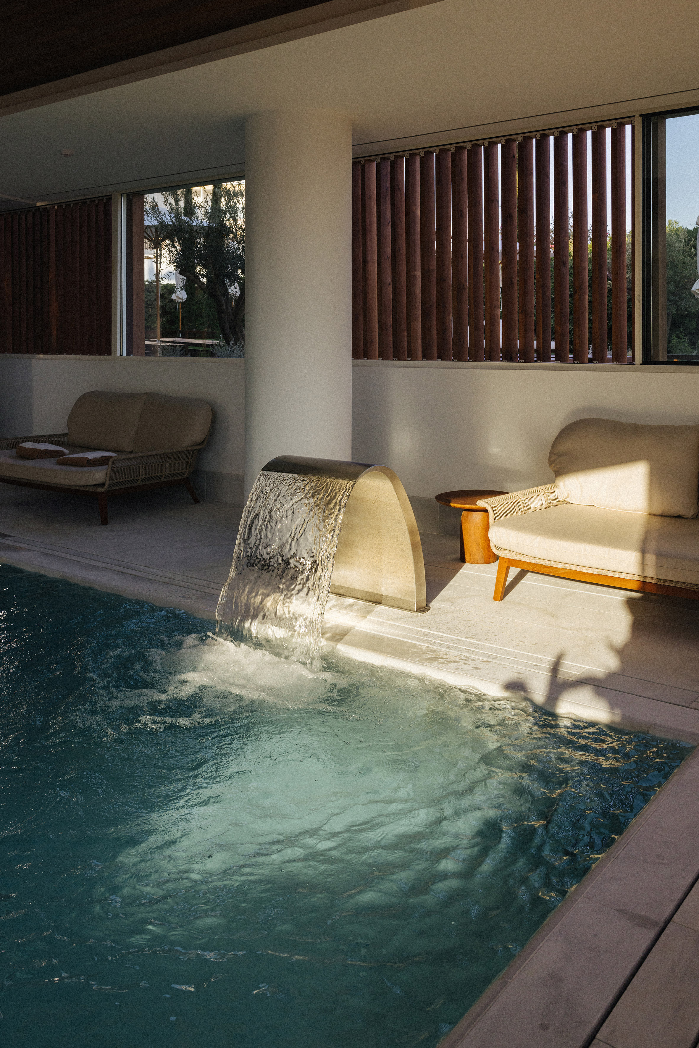 Luxurious indoor pool with waterfall, sunlit daybeds, and wooden blinds.