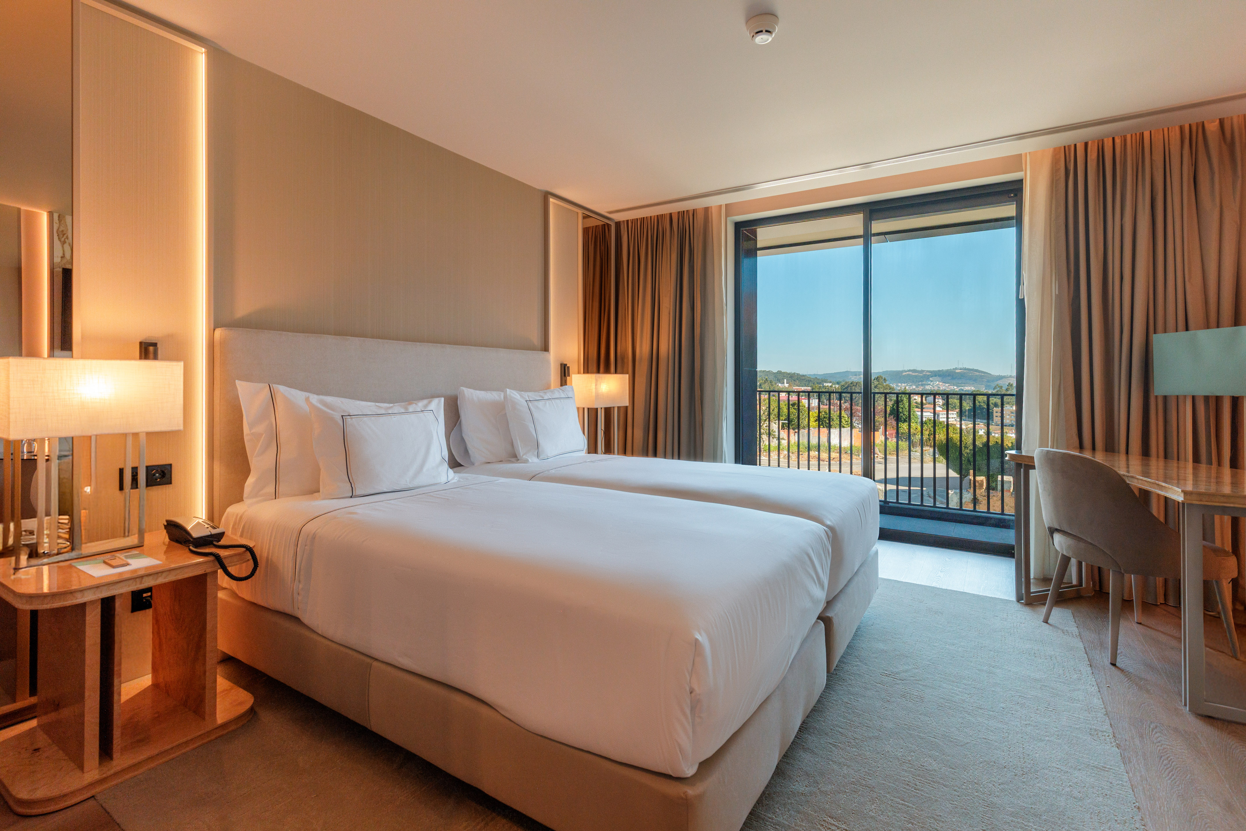 Modern hotel room with twin beds, bedside lamp, and scenic balcony view.