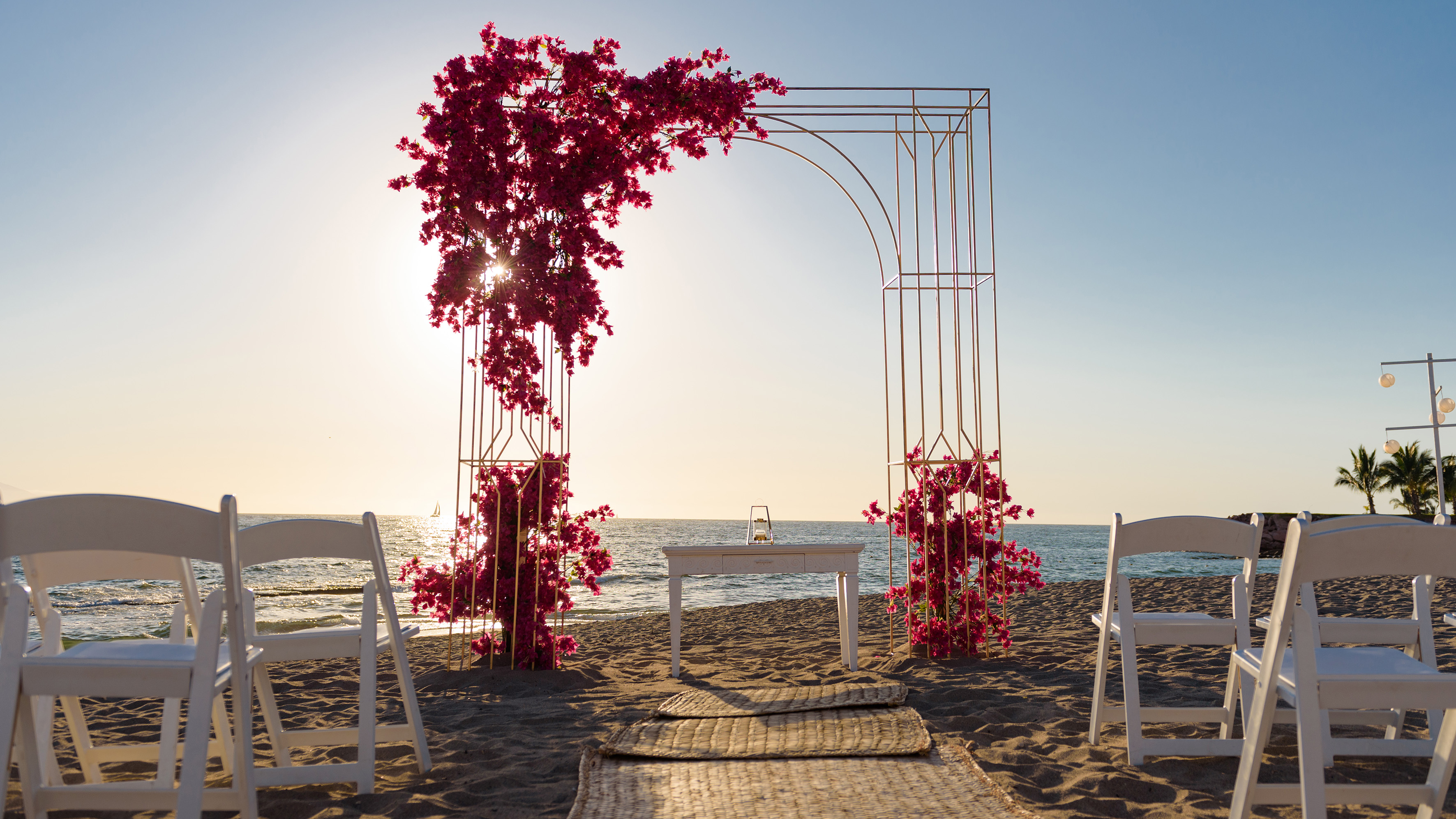 Beach wedding arch with pink bougainvillea, white chairs, and ocean at sunset.