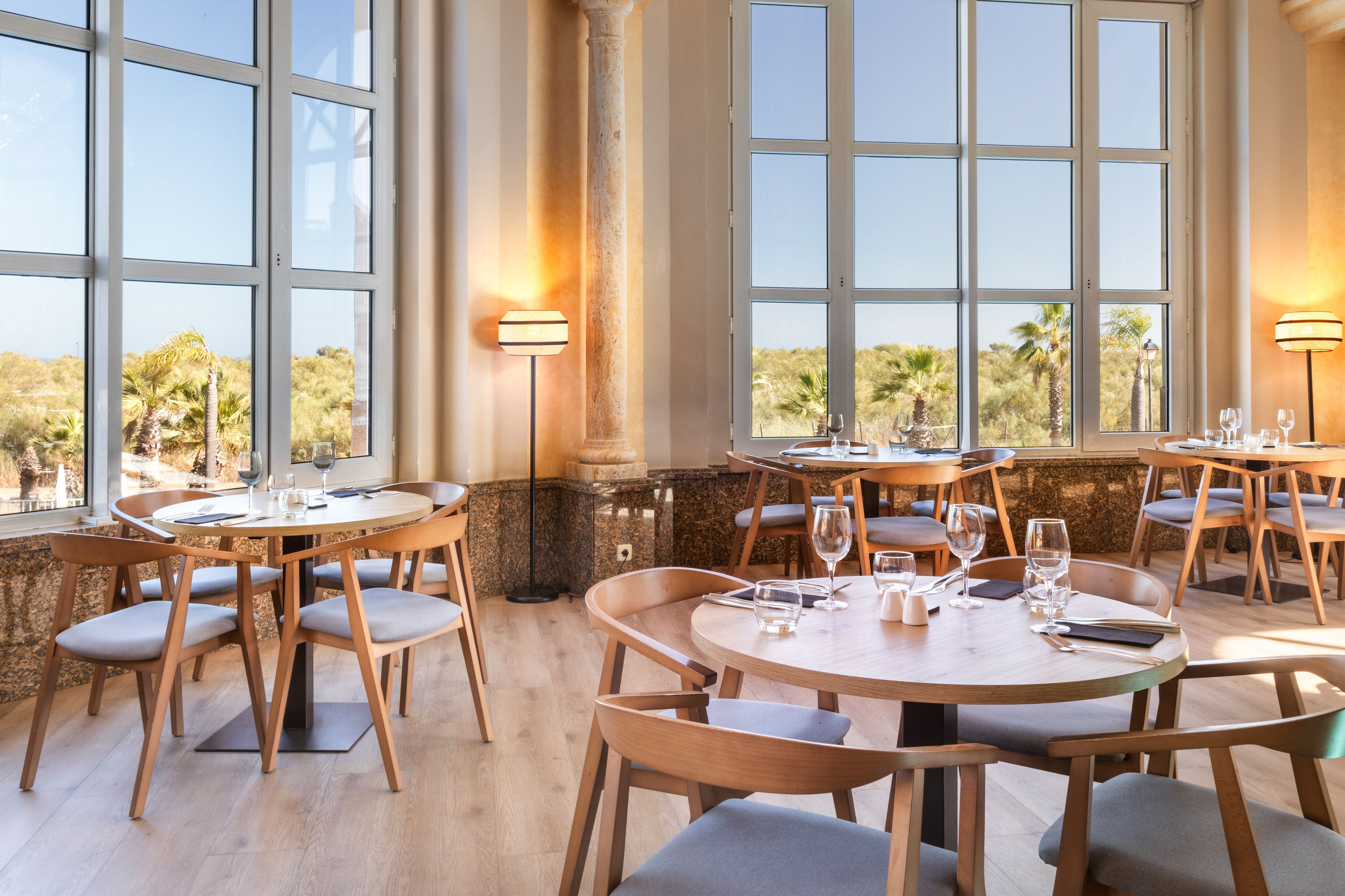Modern restaurant dining room with tables, chairs, large windows, and sunny palm view.
