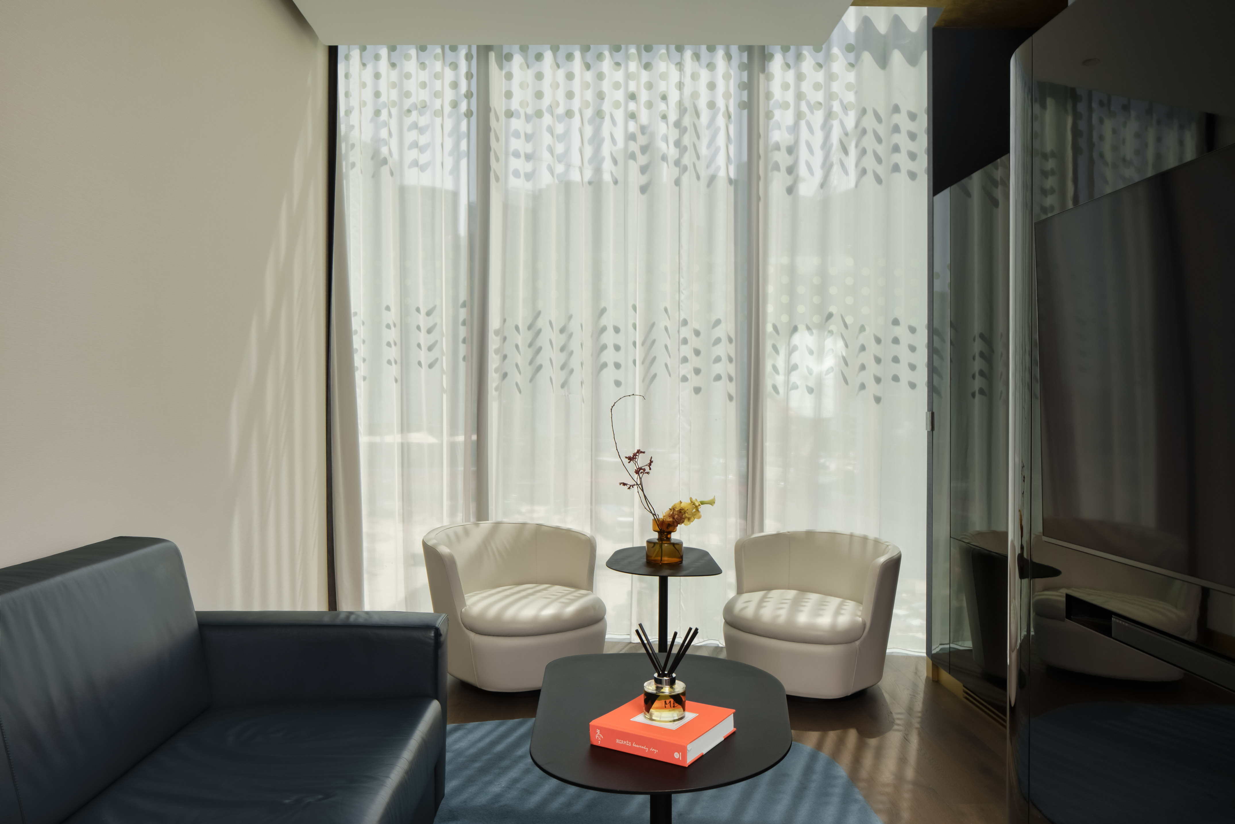 Modern lounge with dark sofa, white armchairs, coffee tables, patterned sheer curtains.