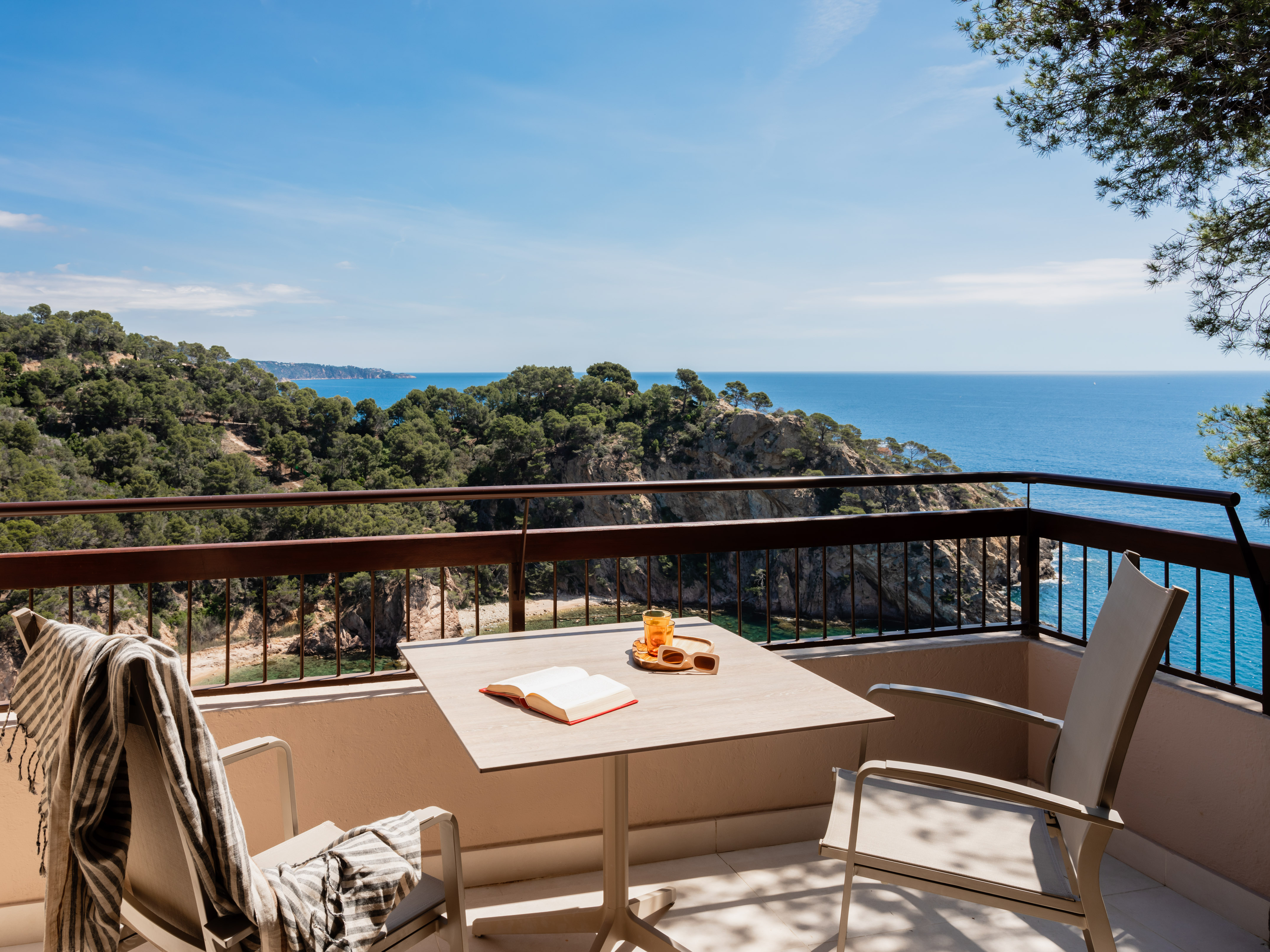 Balcony with open book, drink, chairs, overlooking blue sea and green coast.