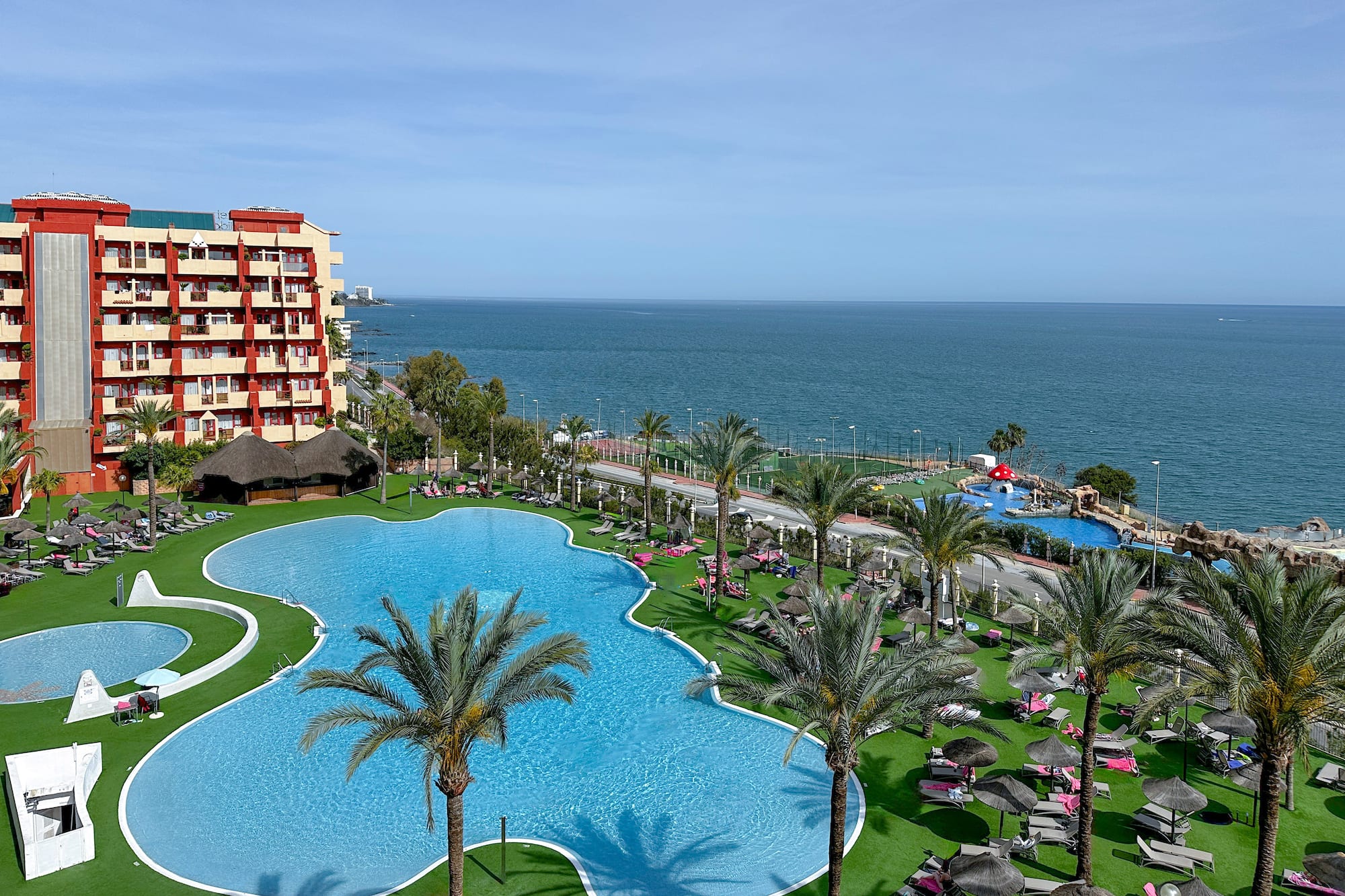 Resort con piscinas, palmeras, tumbonas y hotel junto al mar.