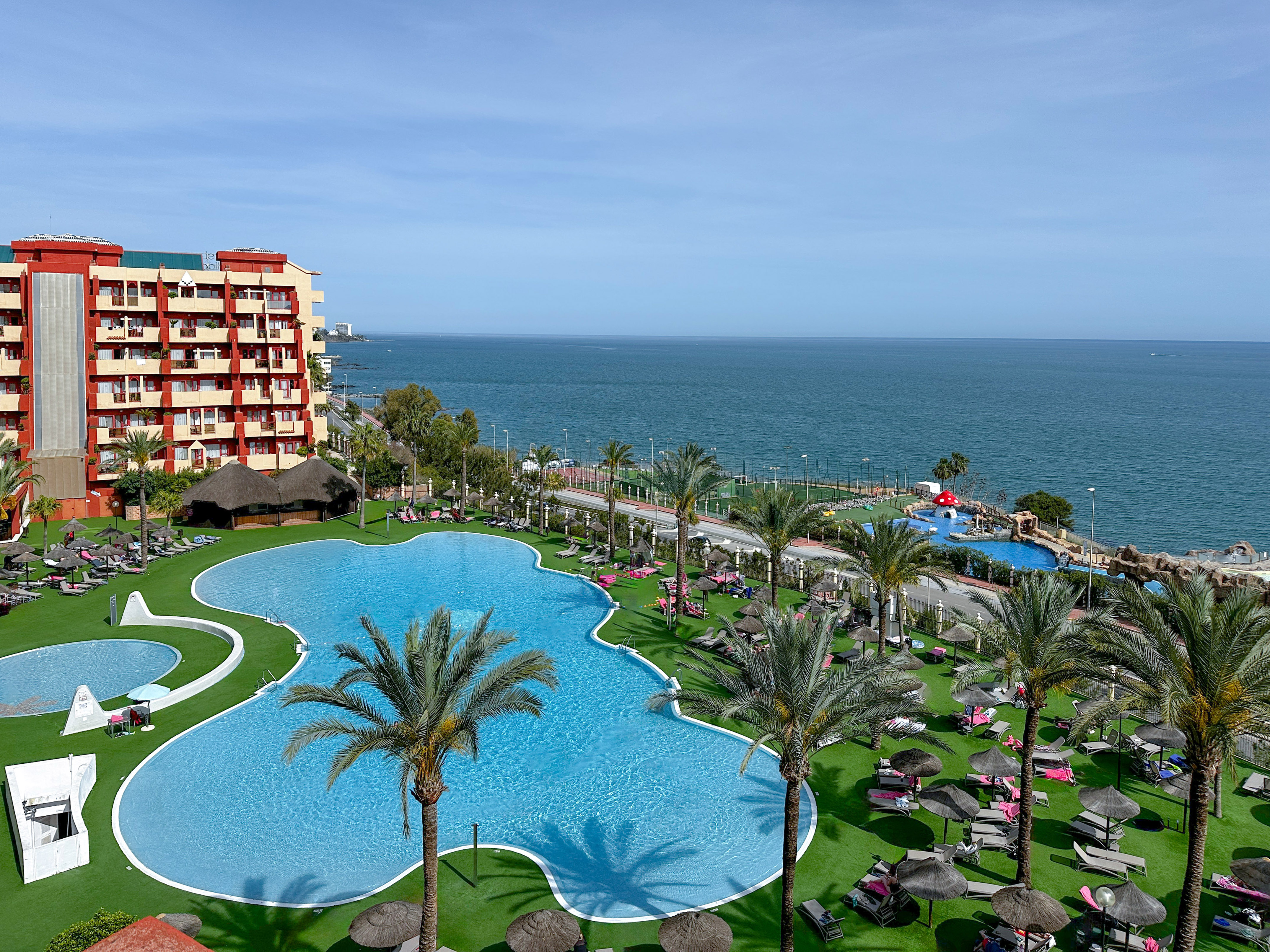 Resort con piscinas, palmeras, tumbonas y hotel junto al mar.