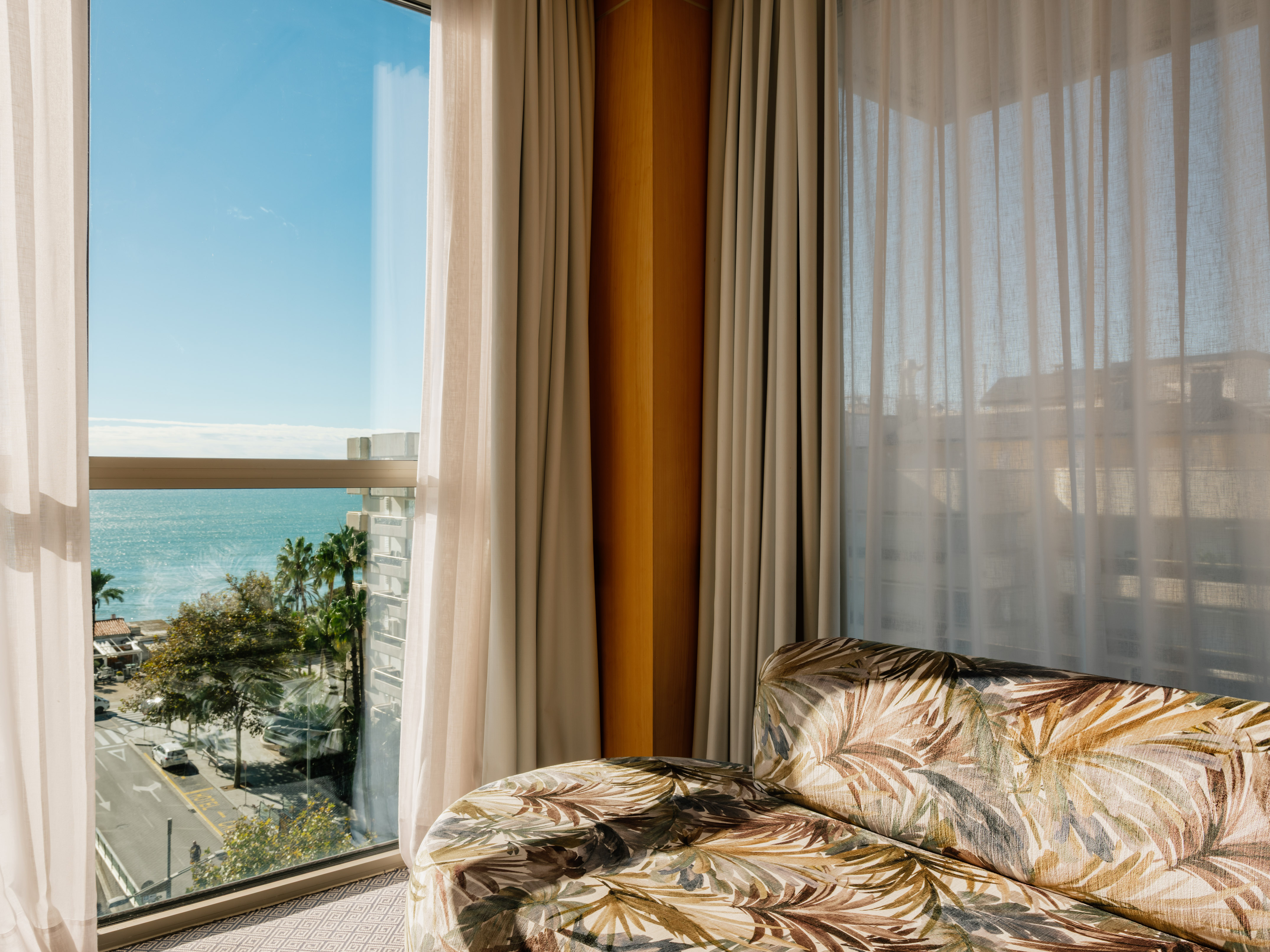 Hotel room with ocean view, street, palm trees, and tropical patterned sofa.