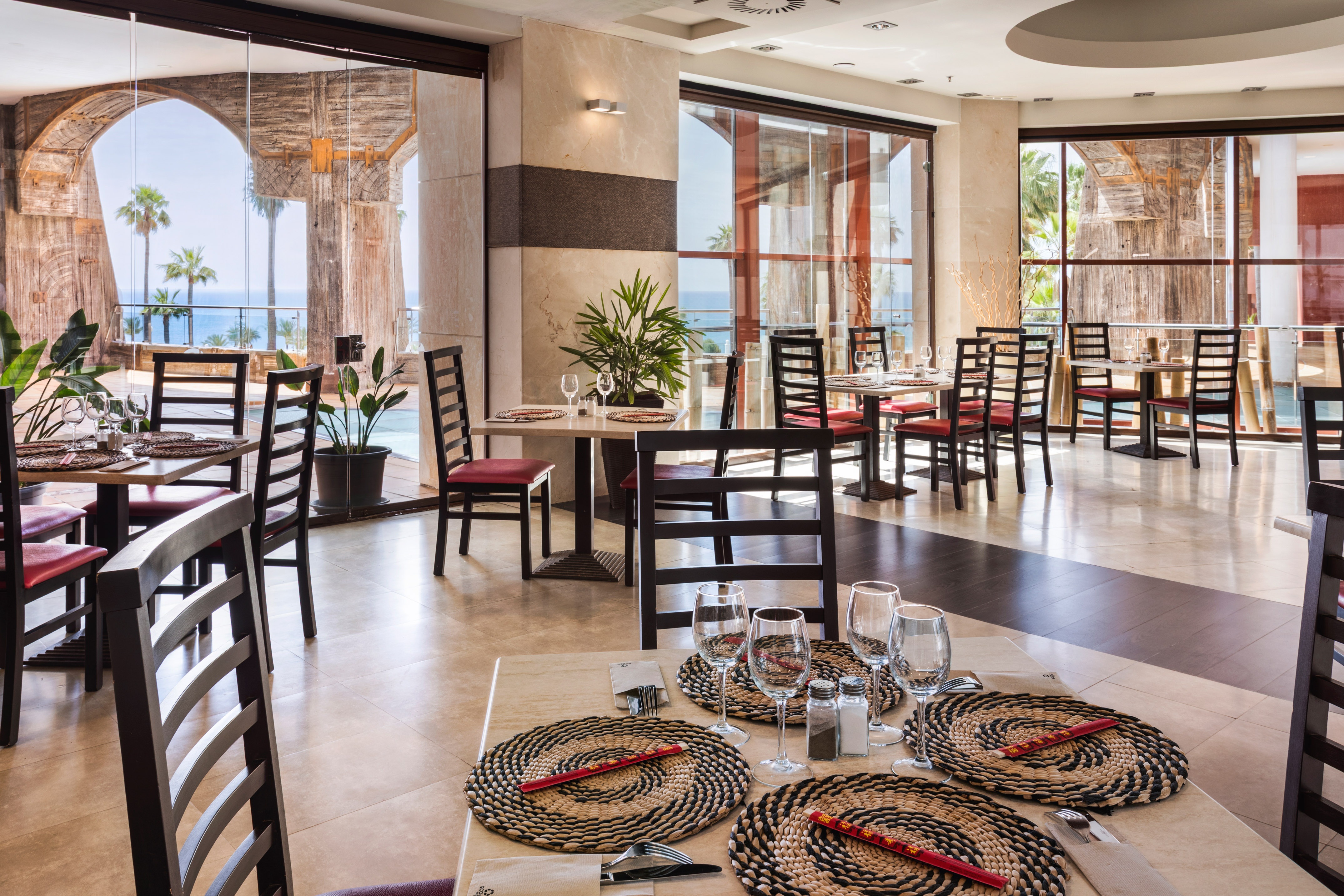 Resort restaurant with ocean view, palm trees, and elegantly set dining tables.