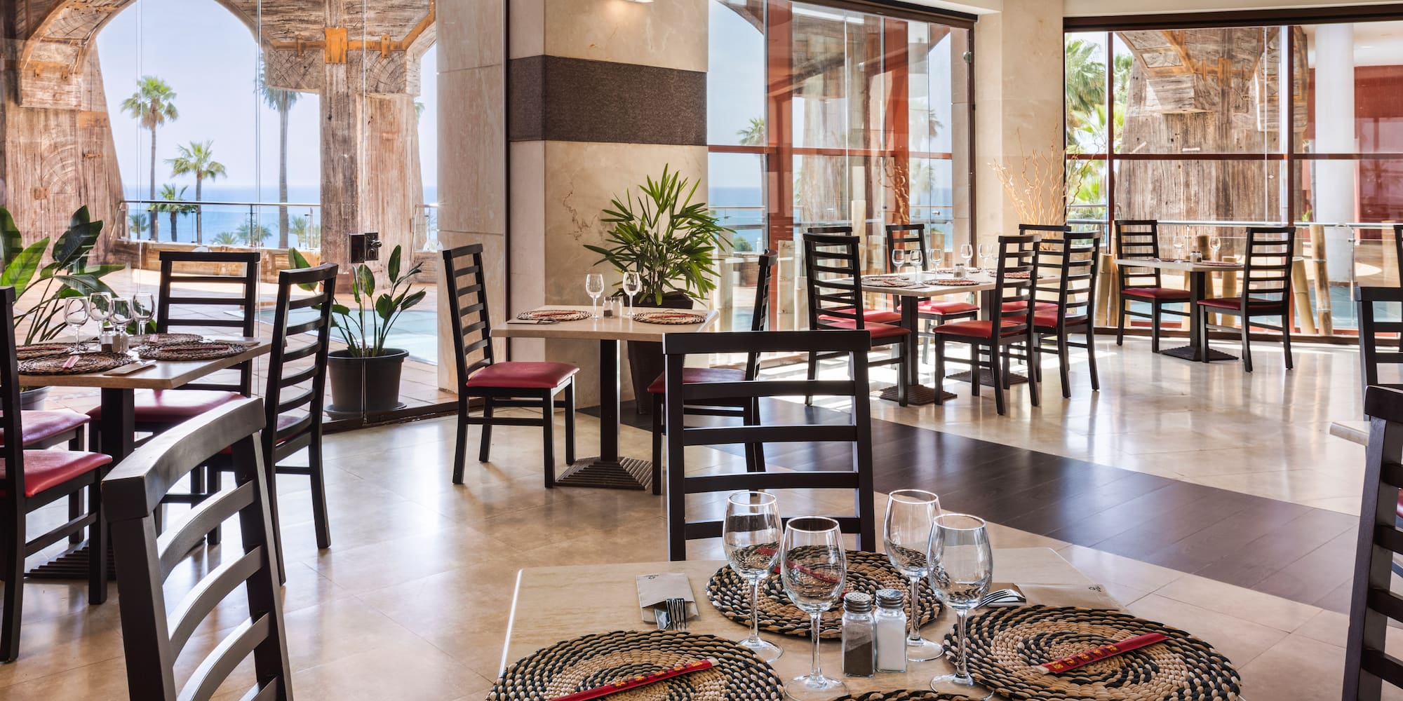 Restaurant interior with ocean view, palm trees, arches, and set tables.