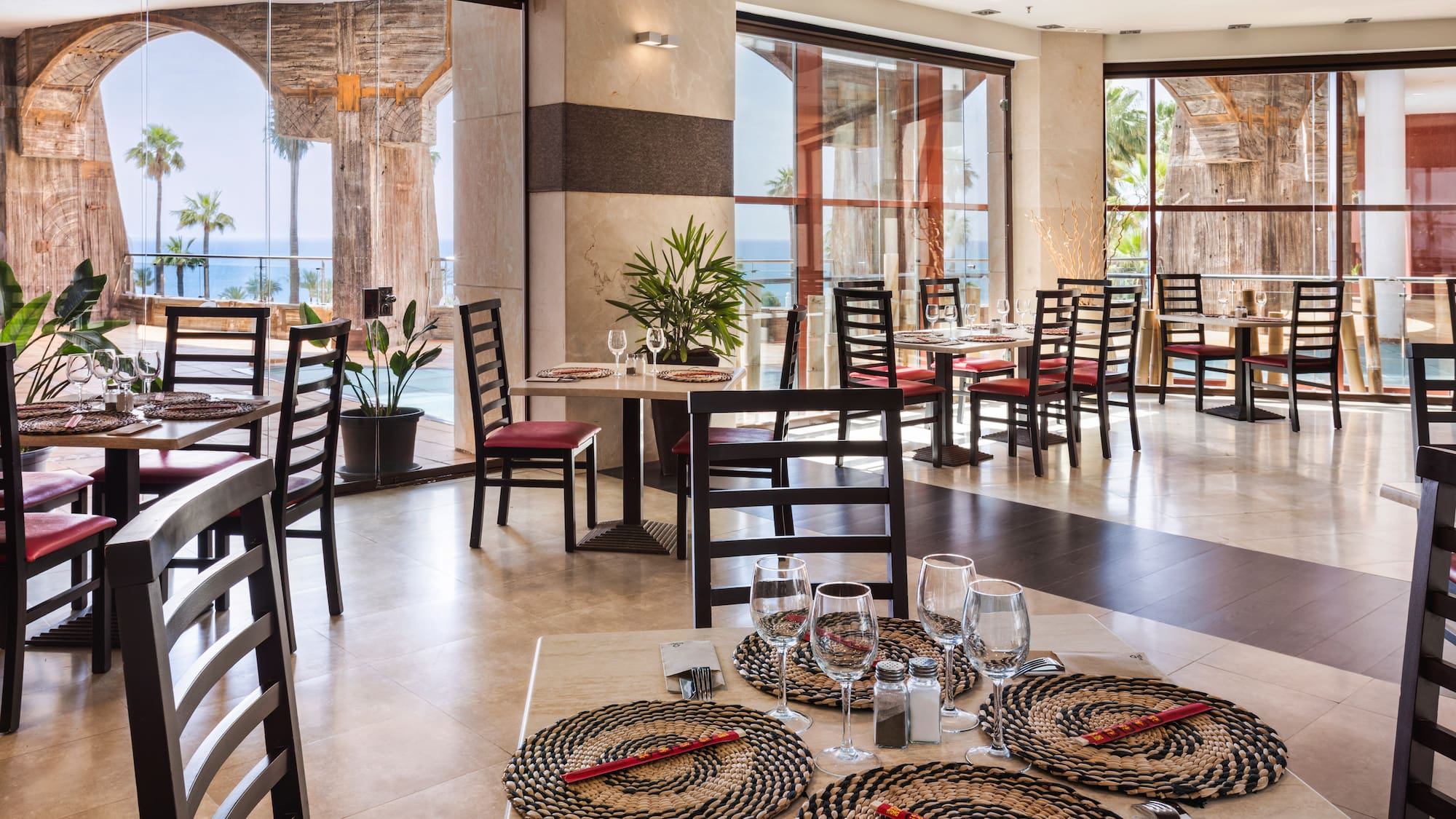 Restaurant interior with ocean view, palm trees, arches, and set tables.