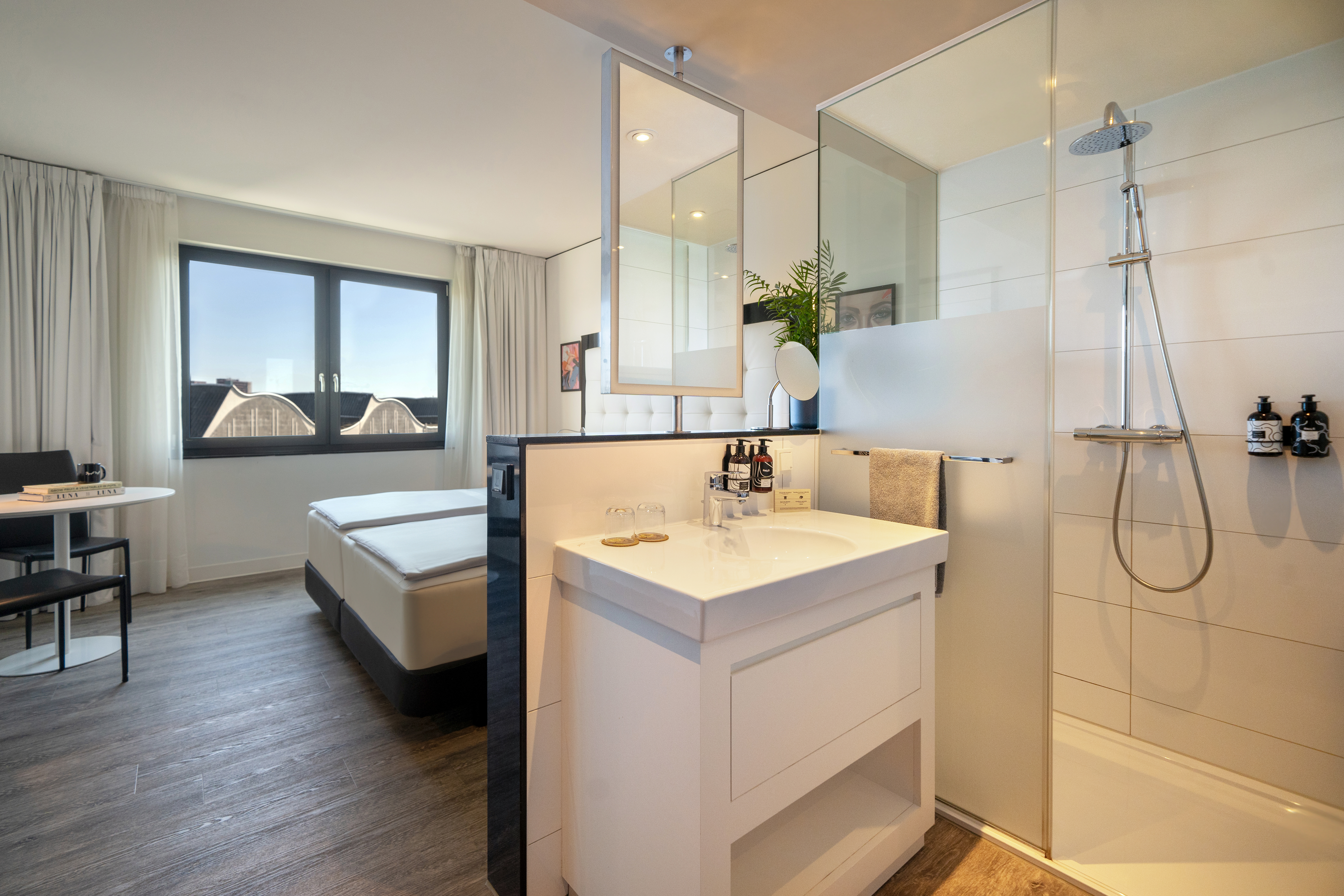 Modern hotel room interior with bed, integrated shower, sink, and window view.