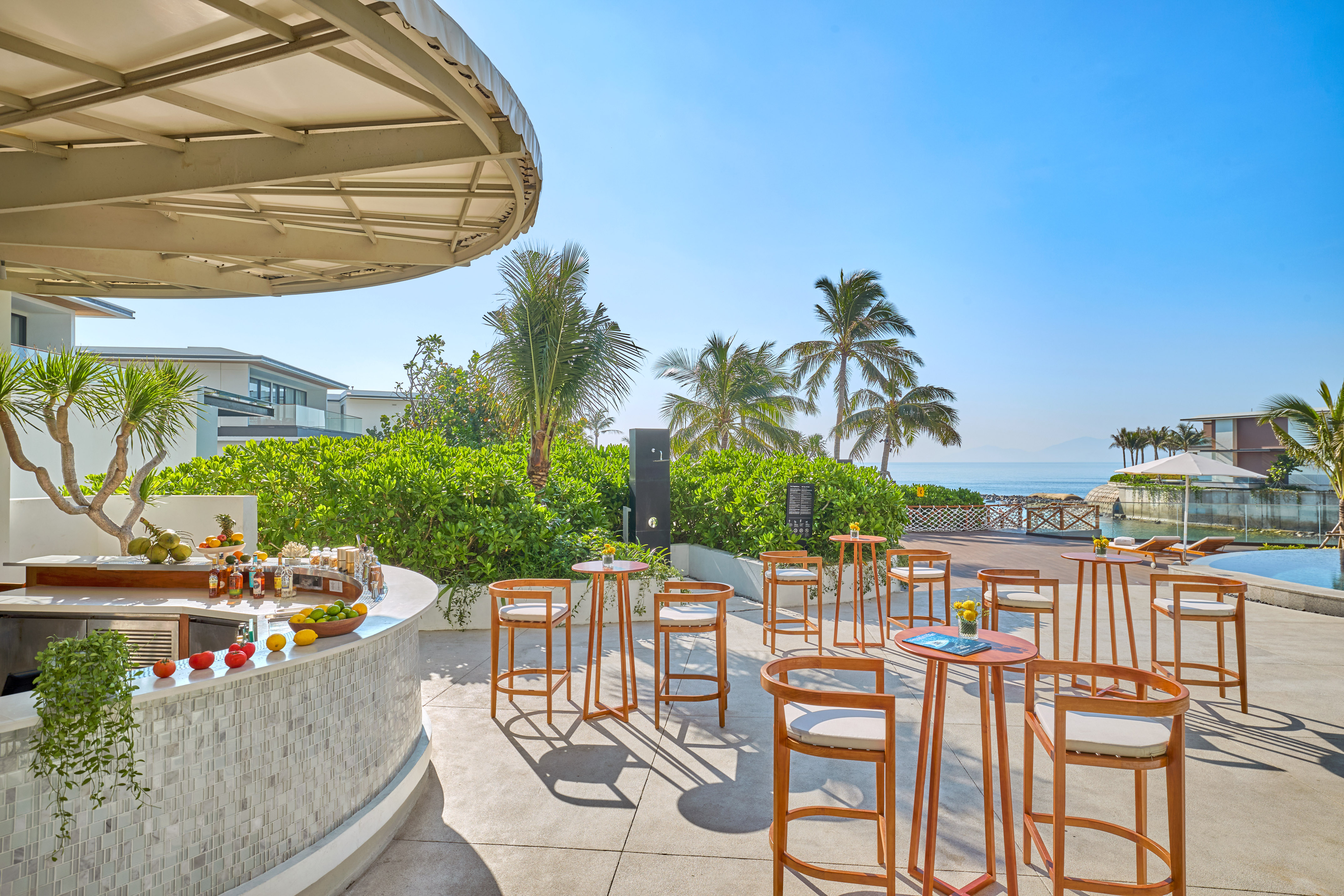 Beachfront resort bar with high tables, lush plants, and ocean view.