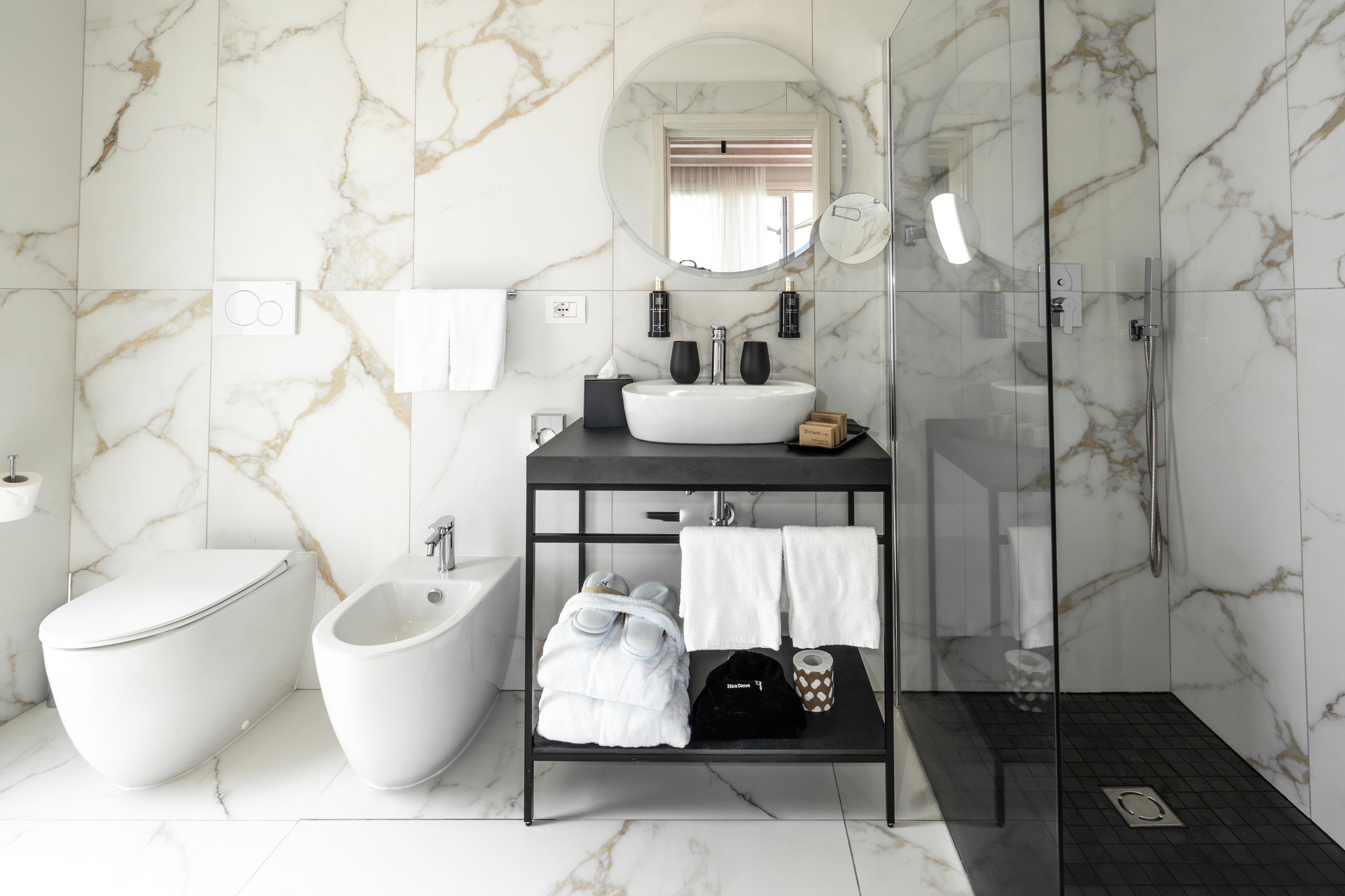 Modern white marble bathroom with black vanity, sink, shower, toilet, and bidet.