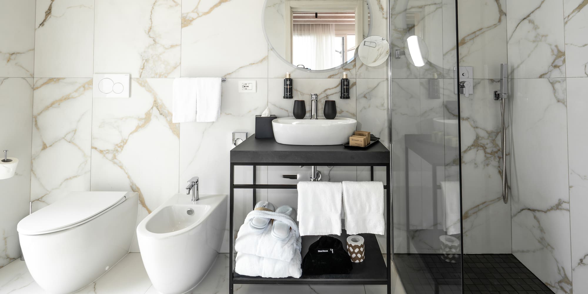 Modern white marble bathroom with black vanity, sink, shower, toilet, and bidet.