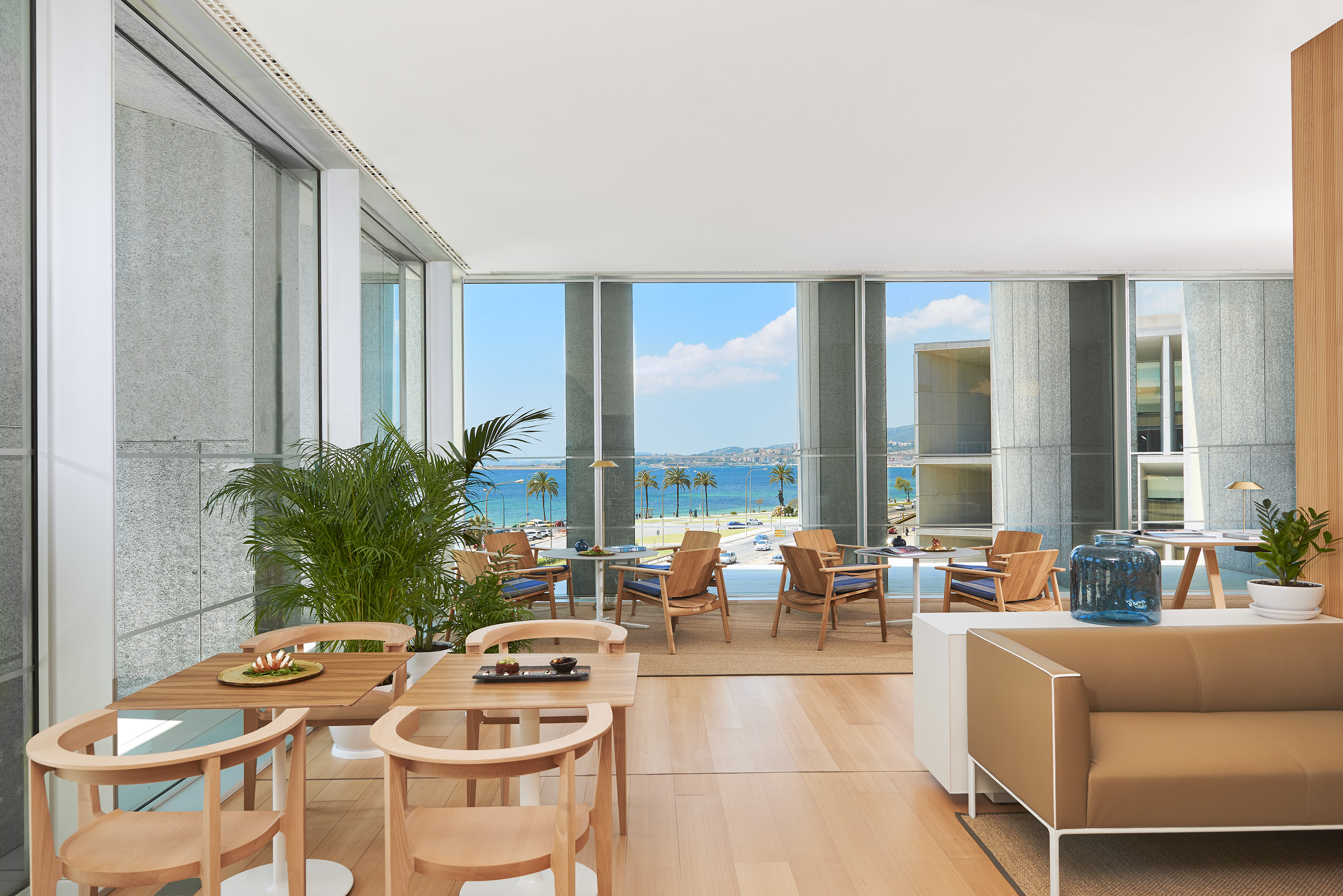 Modern restaurant with ocean view, palm trees, and light wood seating area.