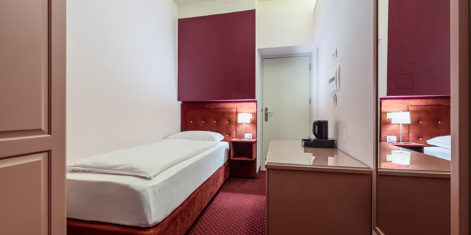 Compact hotel room with single bed, red carpet, bedside lamp, and work desk.