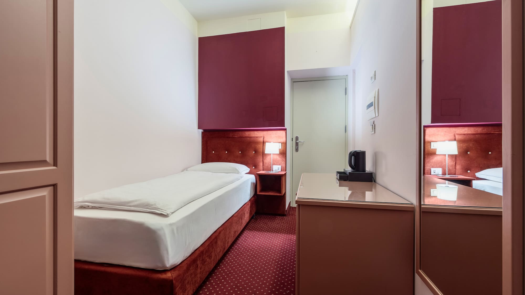 Compact hotel room with single bed, red carpet, and accent wall.