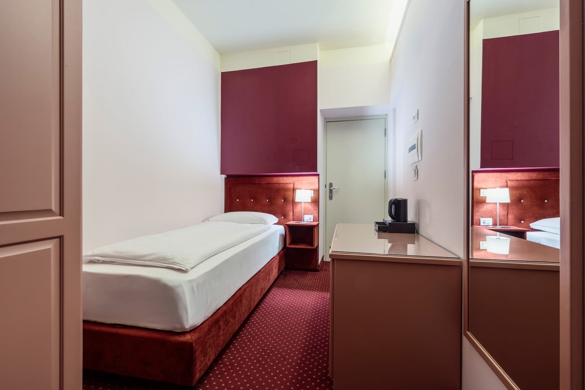Compact hotel room with single bed, red carpet, bedside lamp, and work desk.