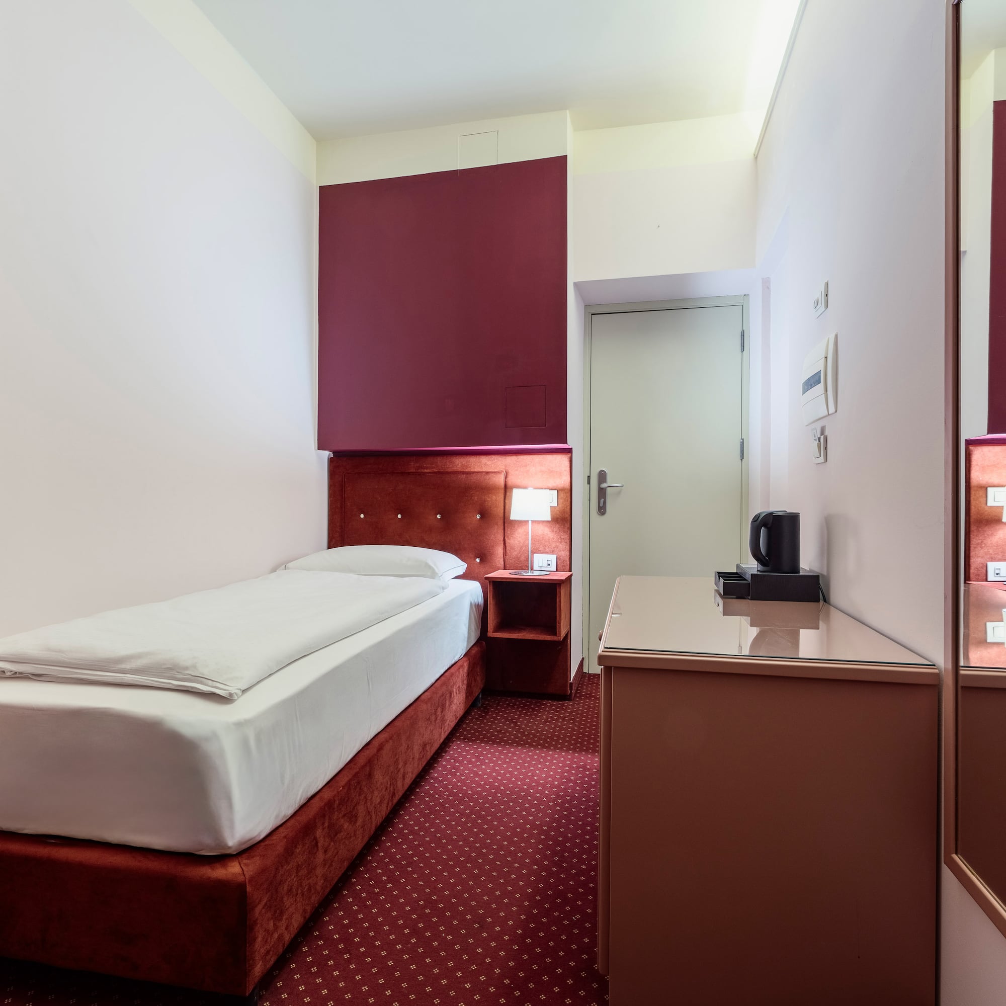 Compact hotel room with single bed, red carpet, bedside lamp, and work desk.