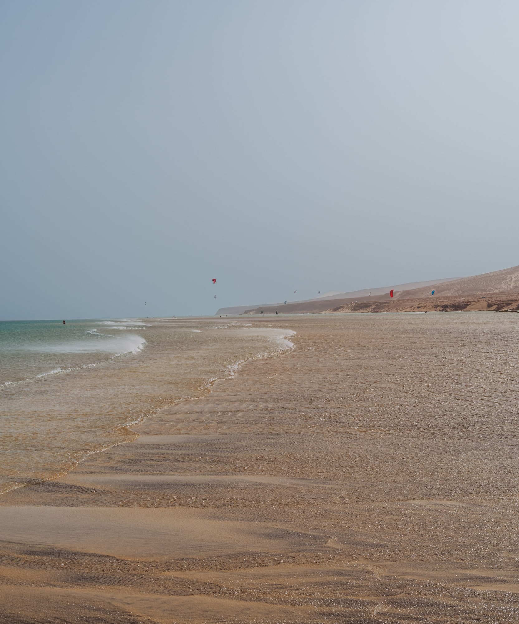 Playa de arena con mar, kitesurfistas y colina desértica.