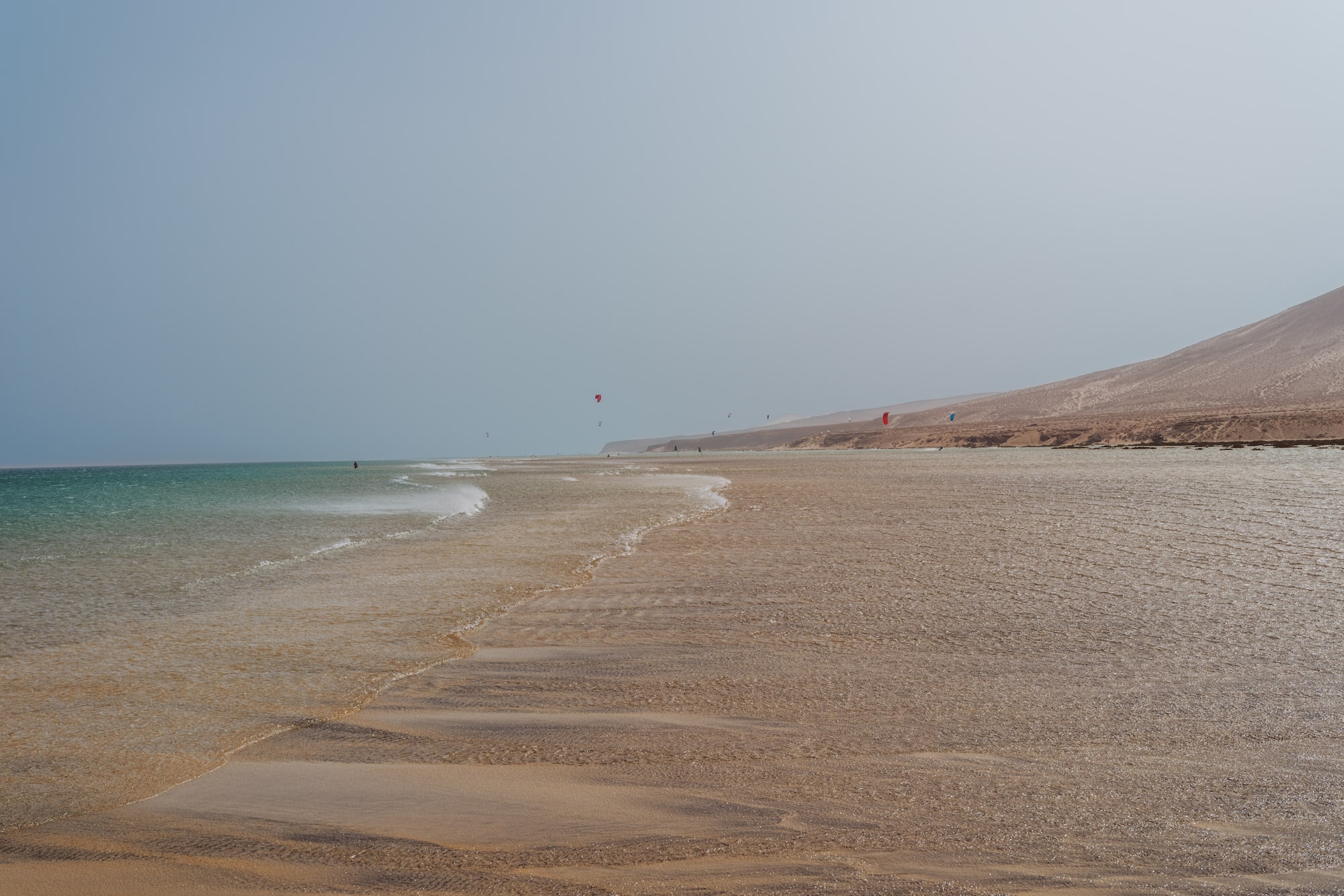 Playa de arena con mar, kitesurfistas y colina desértica.