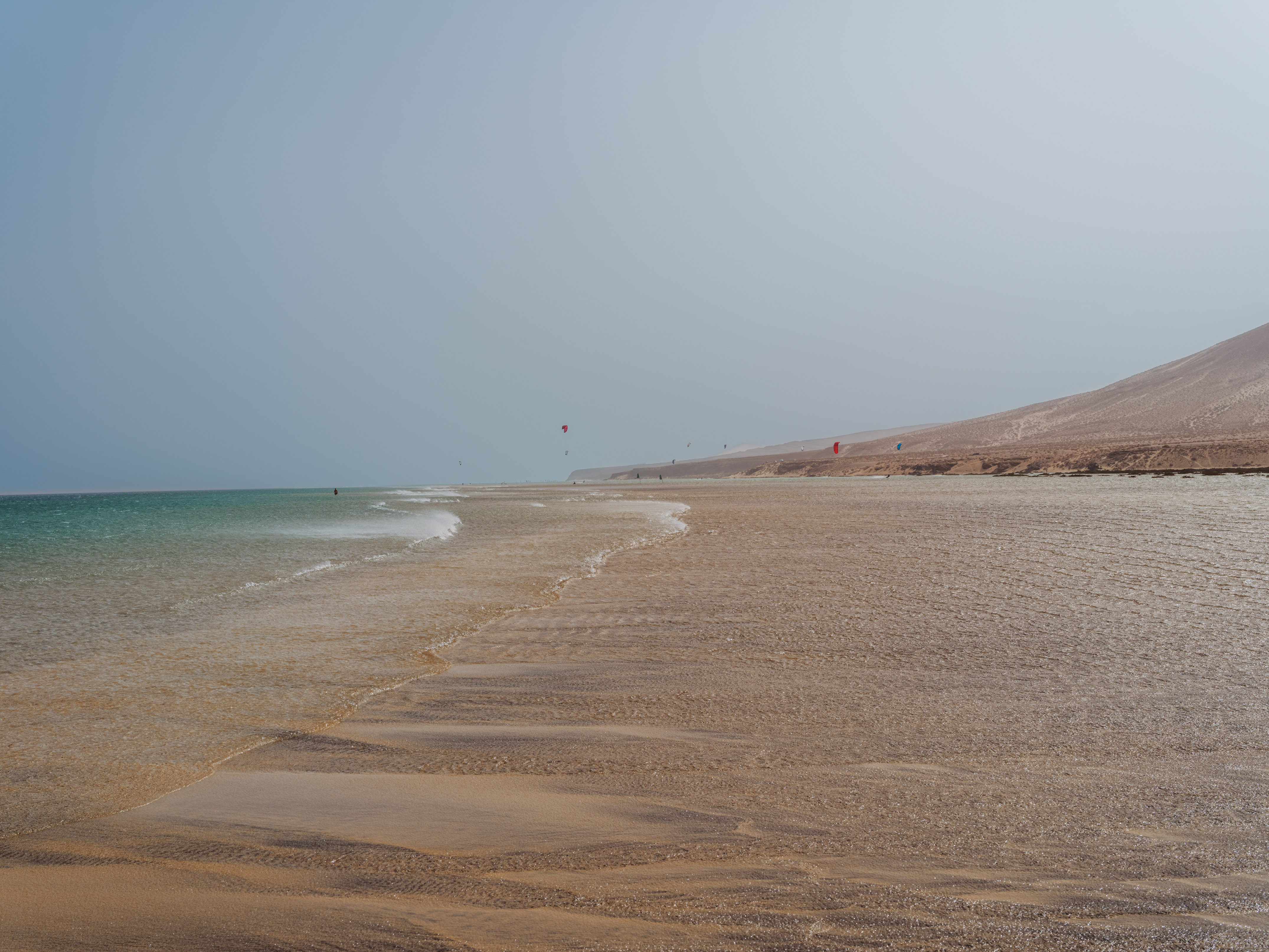 Playa de arena con mar, kitesurfistas y colina desértica.