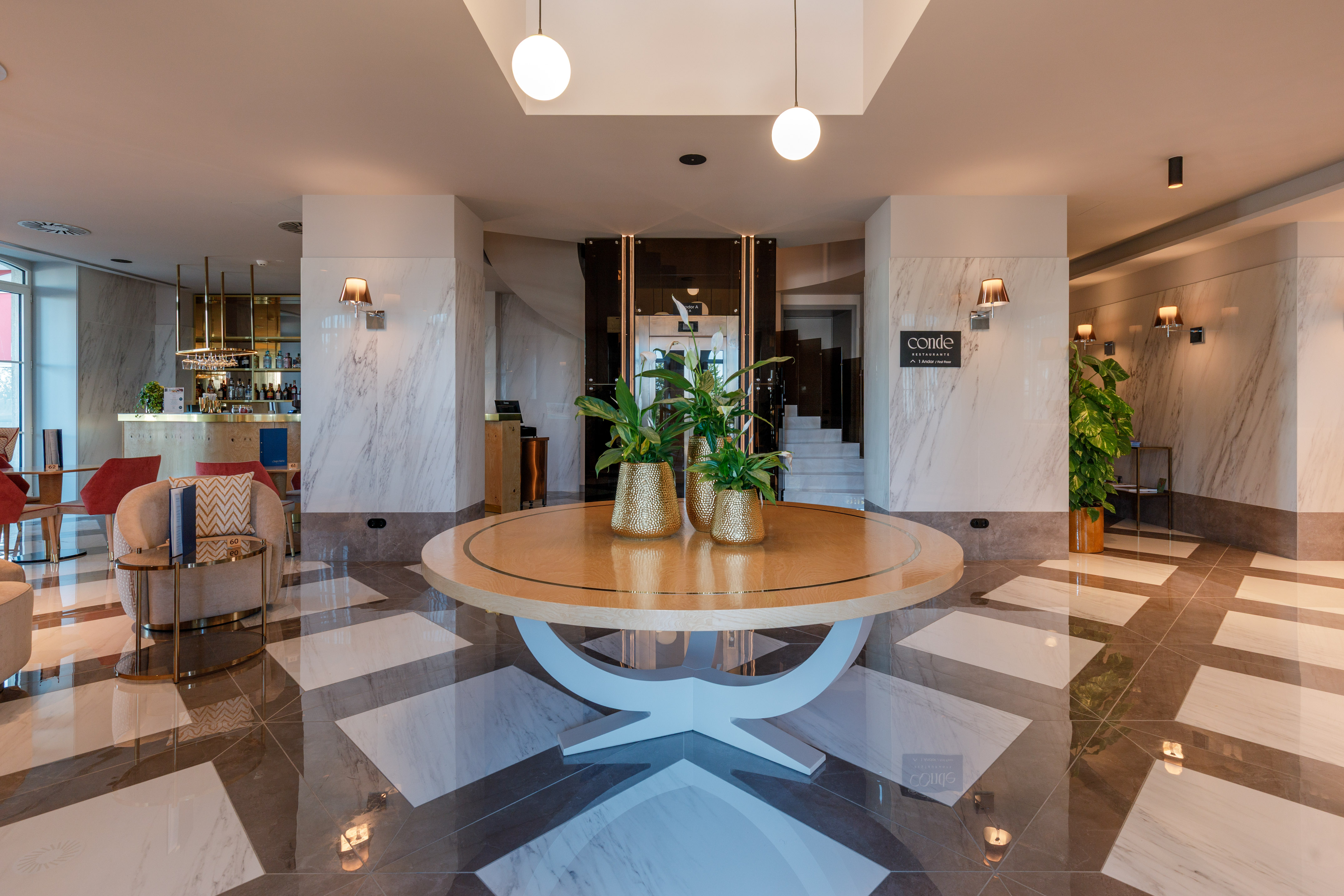 Modern hotel lobby with round table, potted plants, bar, and patterned marble floor.