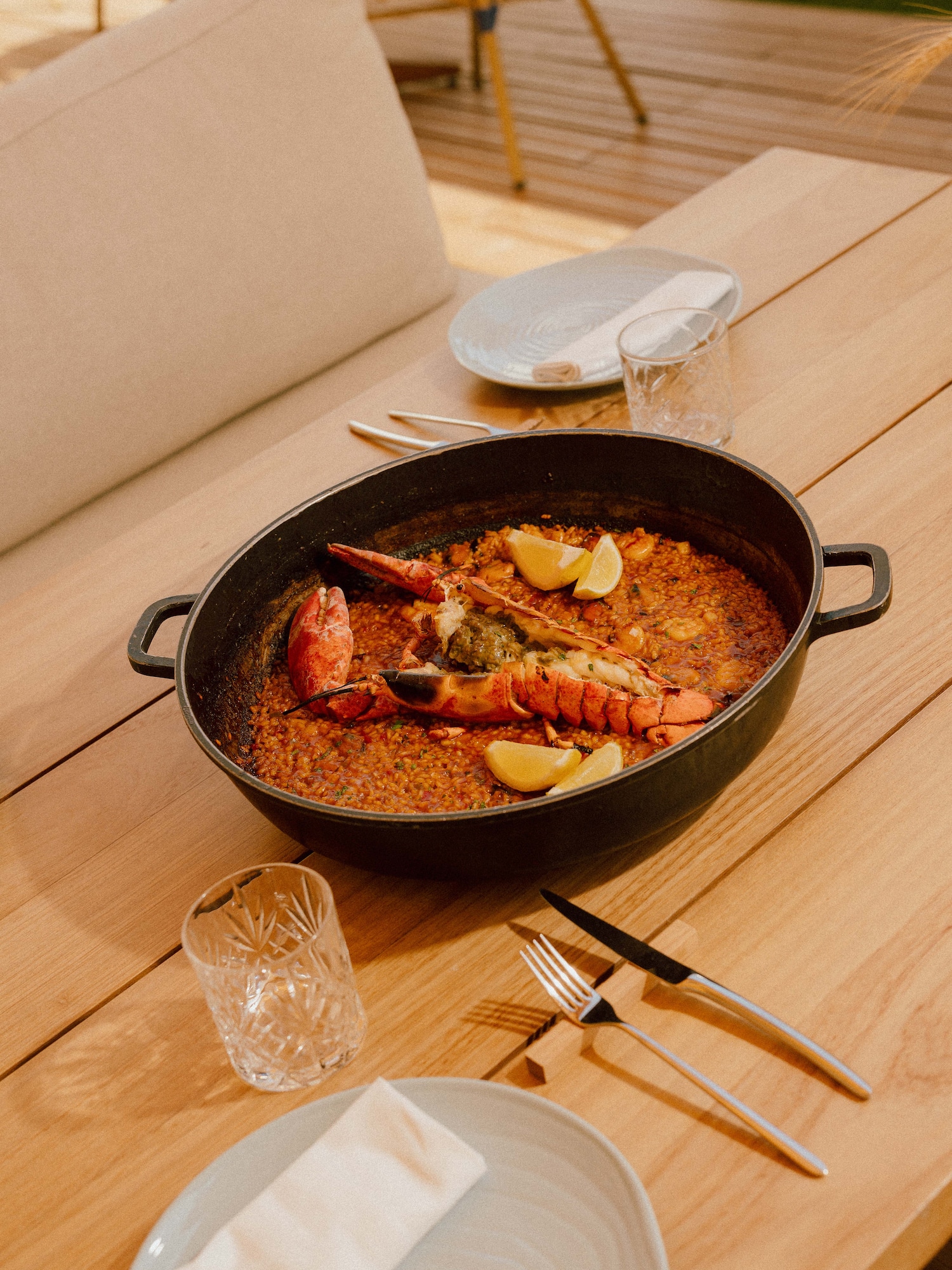 Pan of lobster paella with lemon wedges on an outdoor wooden table setting.