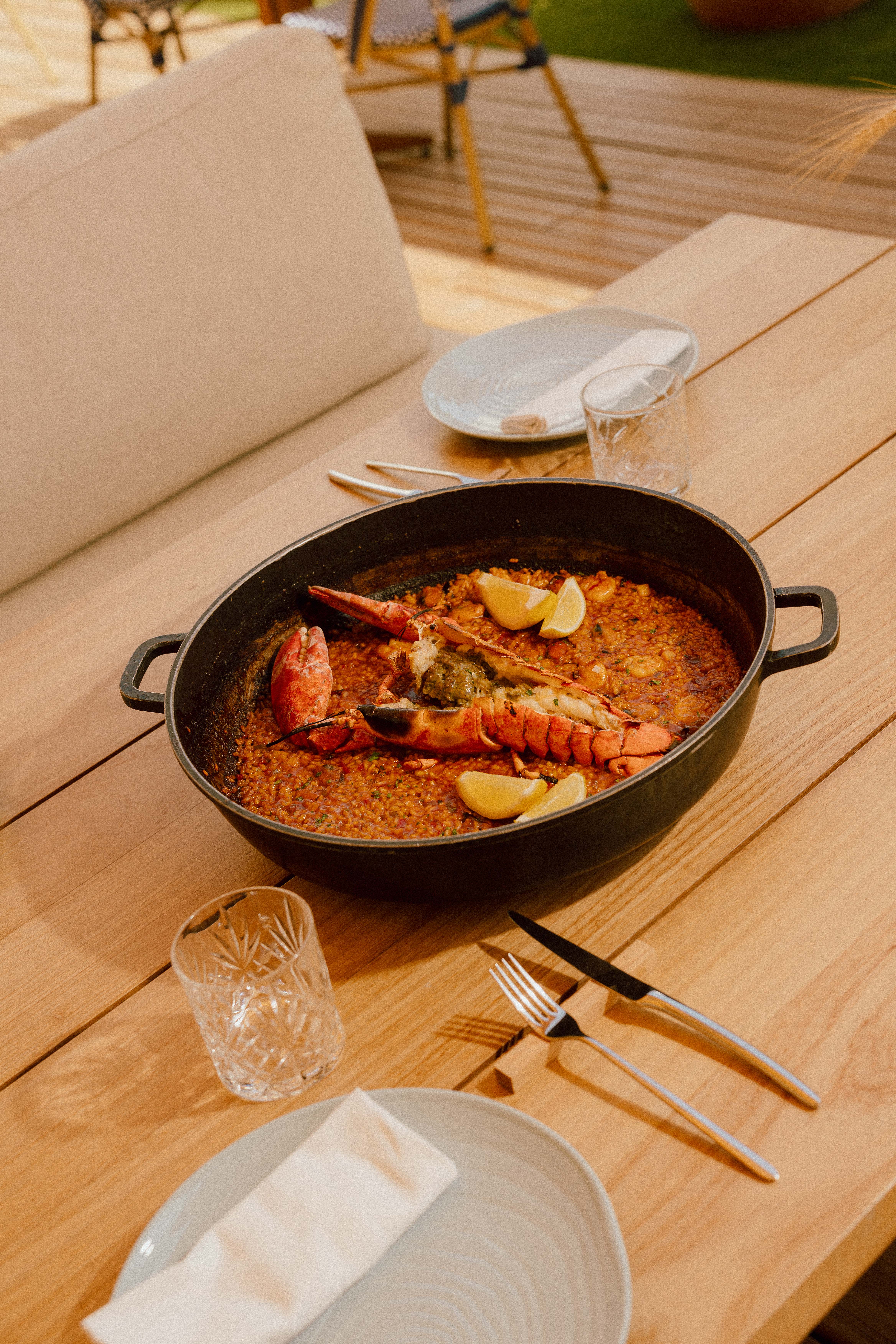 Pan of lobster paella with lemon wedges on an outdoor wooden table setting.