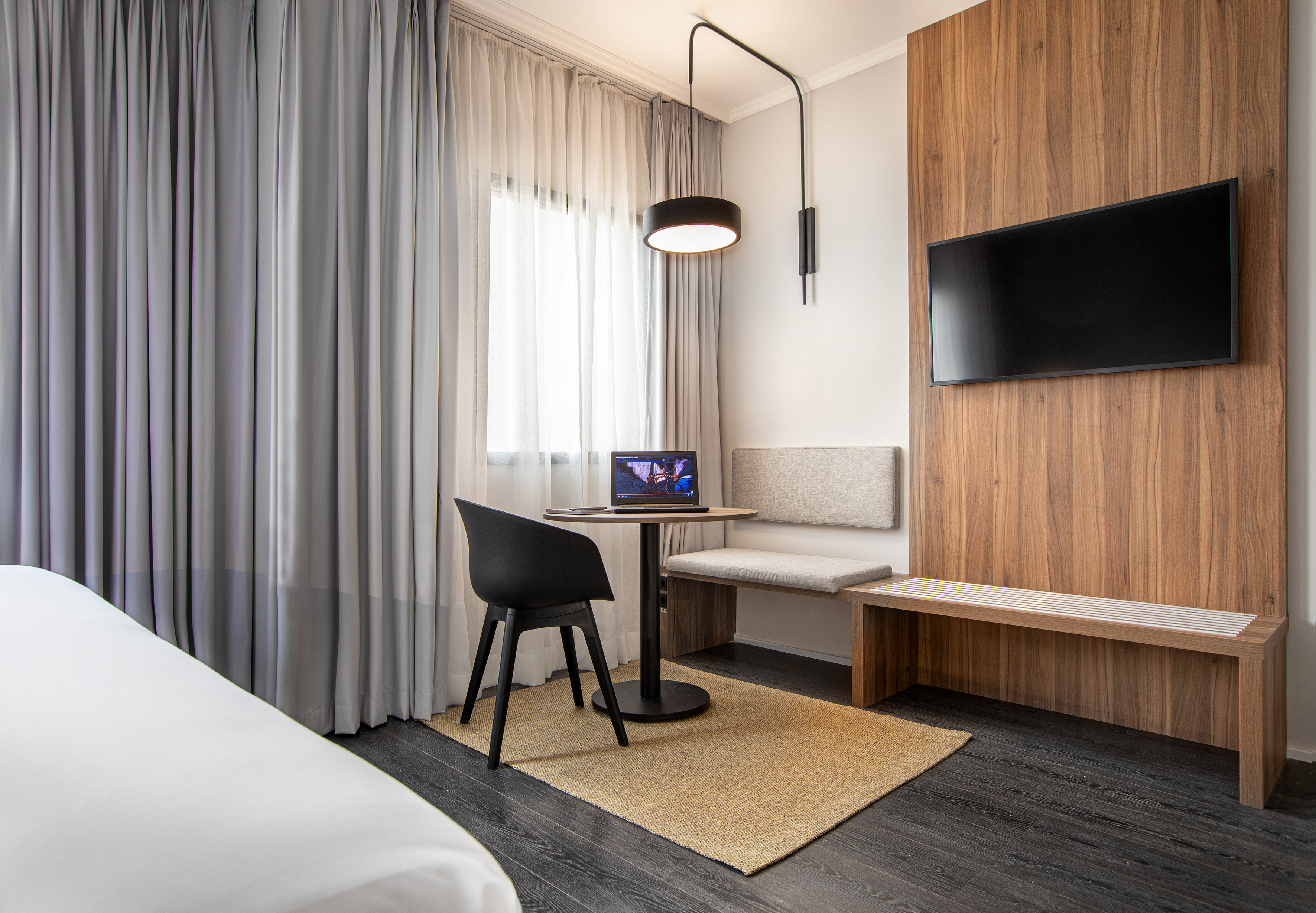Modern guest room with laptop on desk, TV, window, and grey curtains.