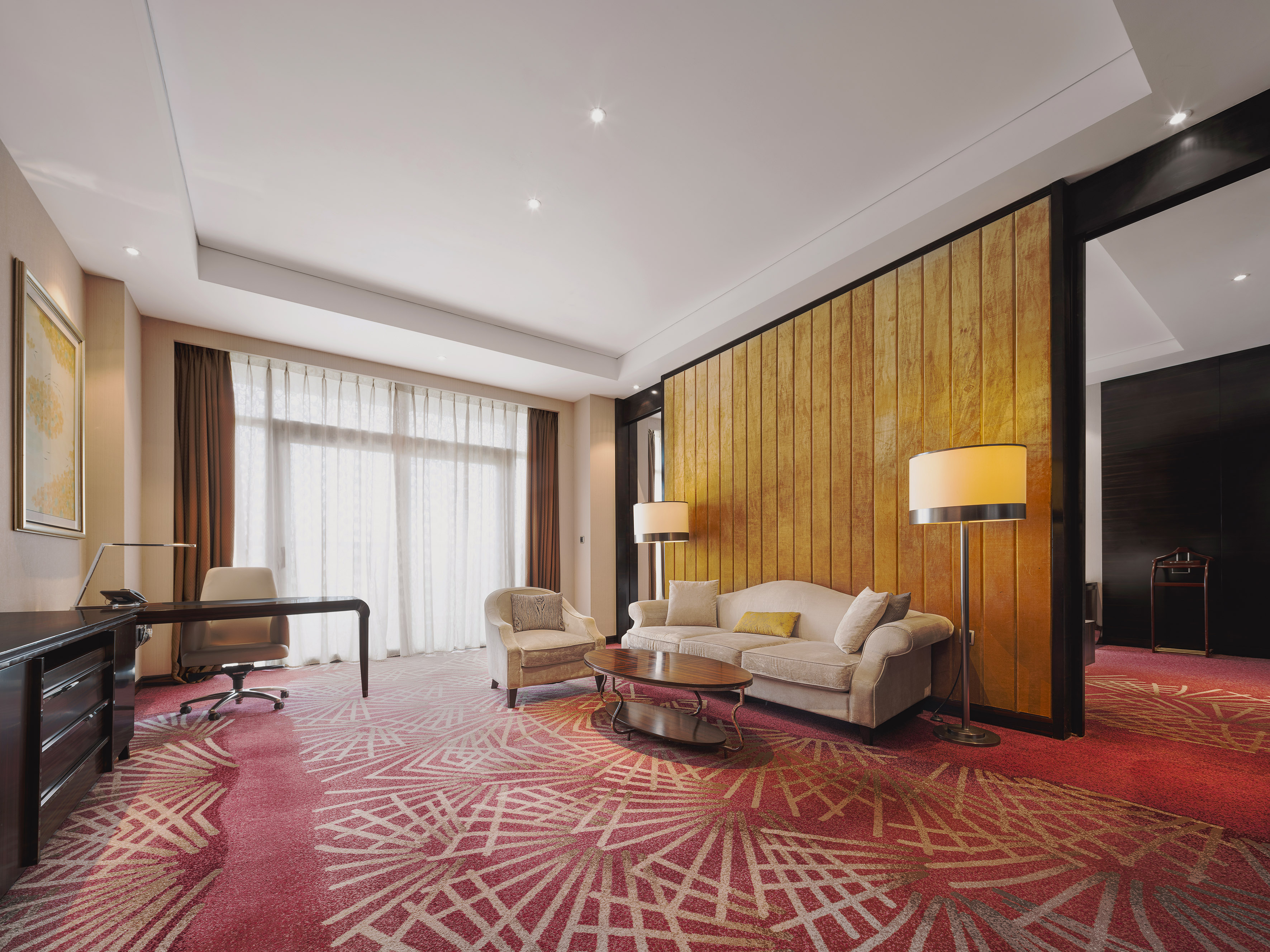 Hotel suite with red geometric carpet, beige sofa, wooden desk, and accent wall.