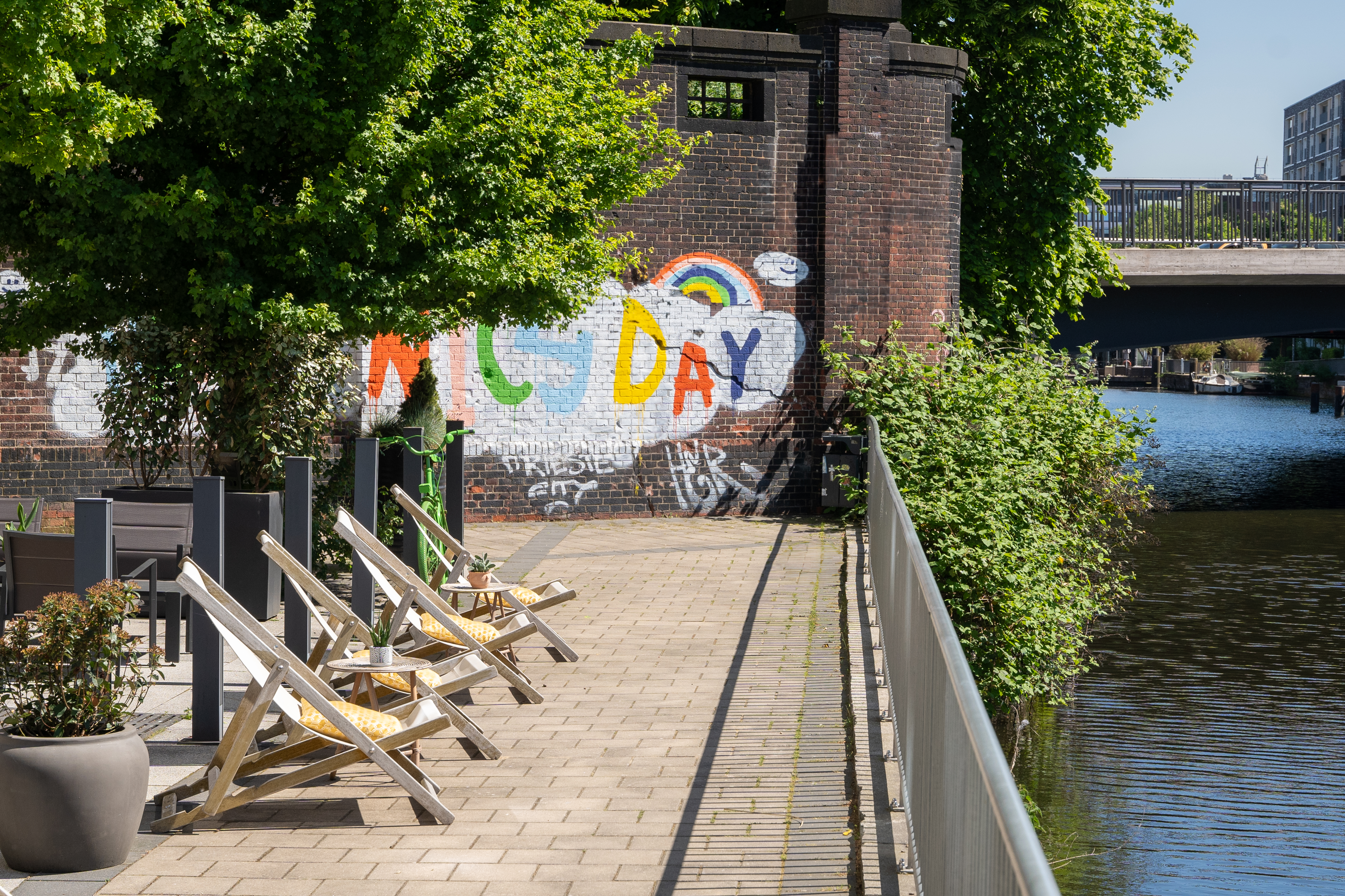 Deck chairs by canal with colorful 'G'DAY' graffiti wall and urban bridge.