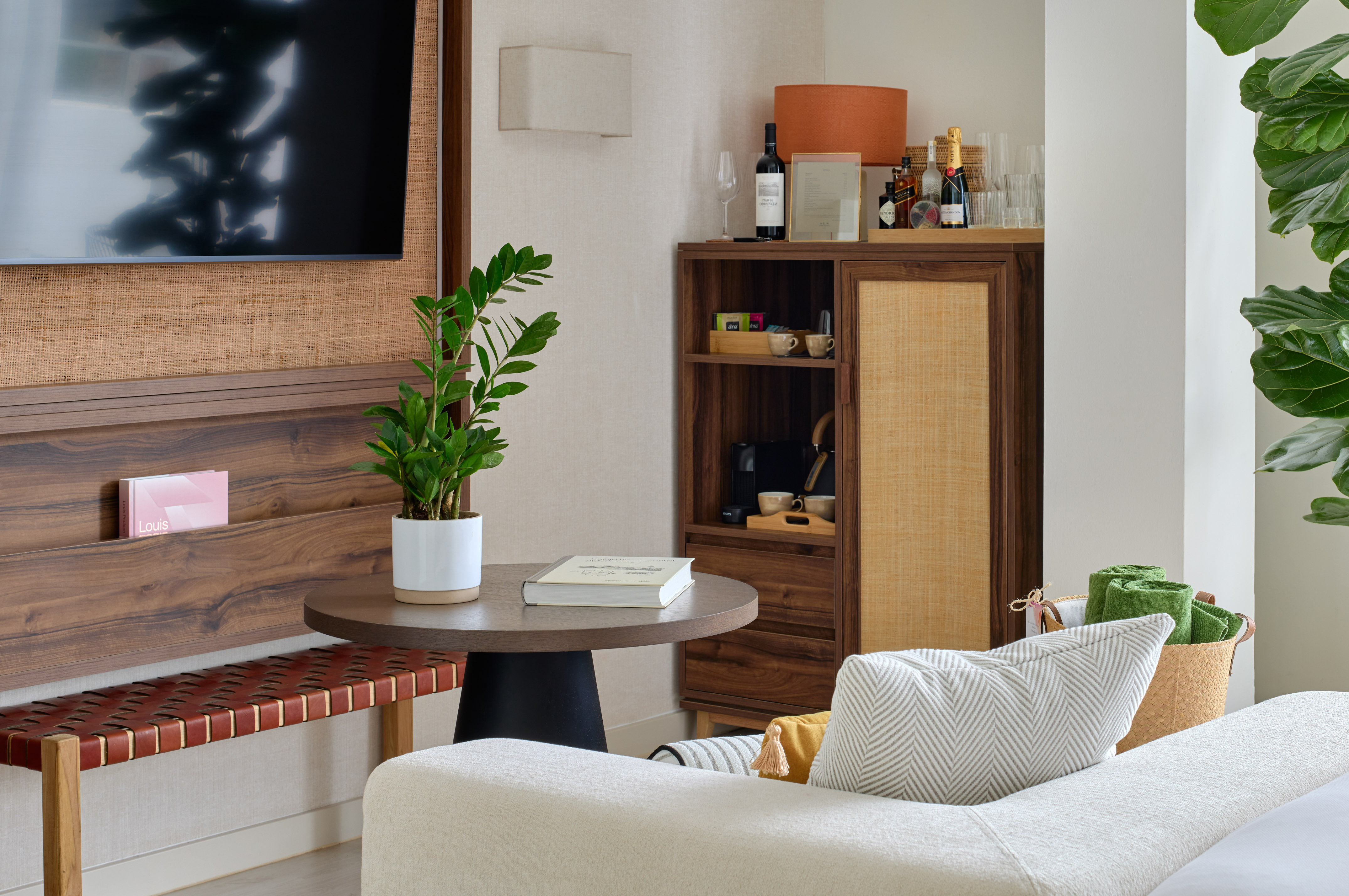 Hotel room lounge with flat-screen TV, wooden minibar, plants, and sofa.