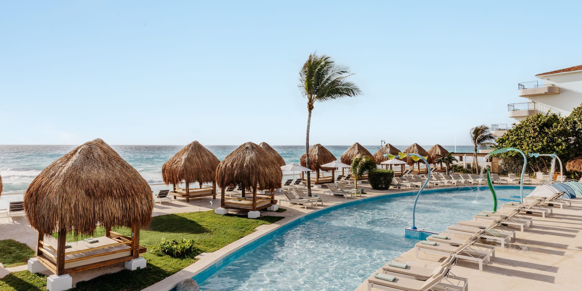 Tropical resort pool with thatched cabanas, ocean, and lounge chairs.