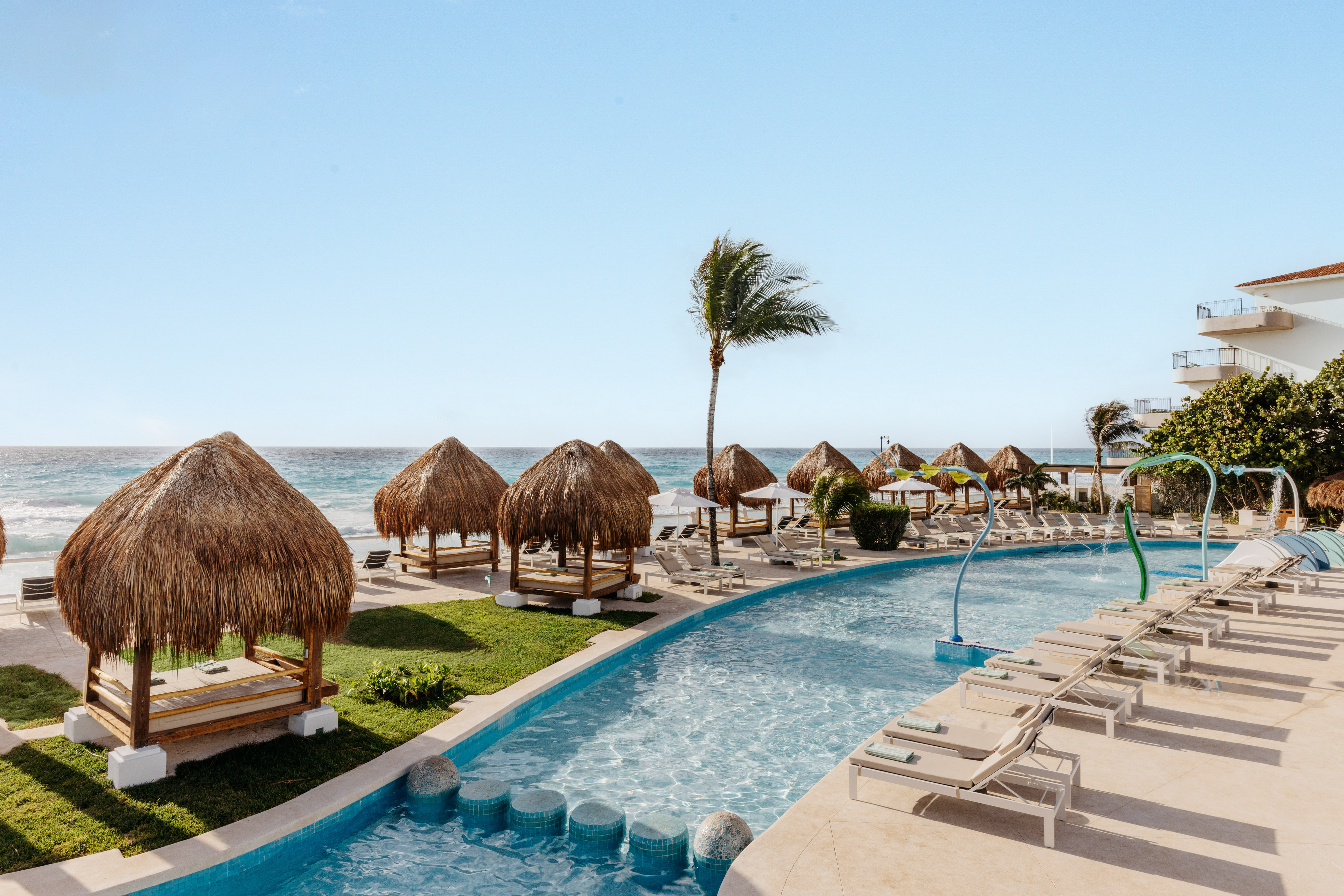 Tropical resort pool with thatched cabanas, ocean, and lounge chairs.