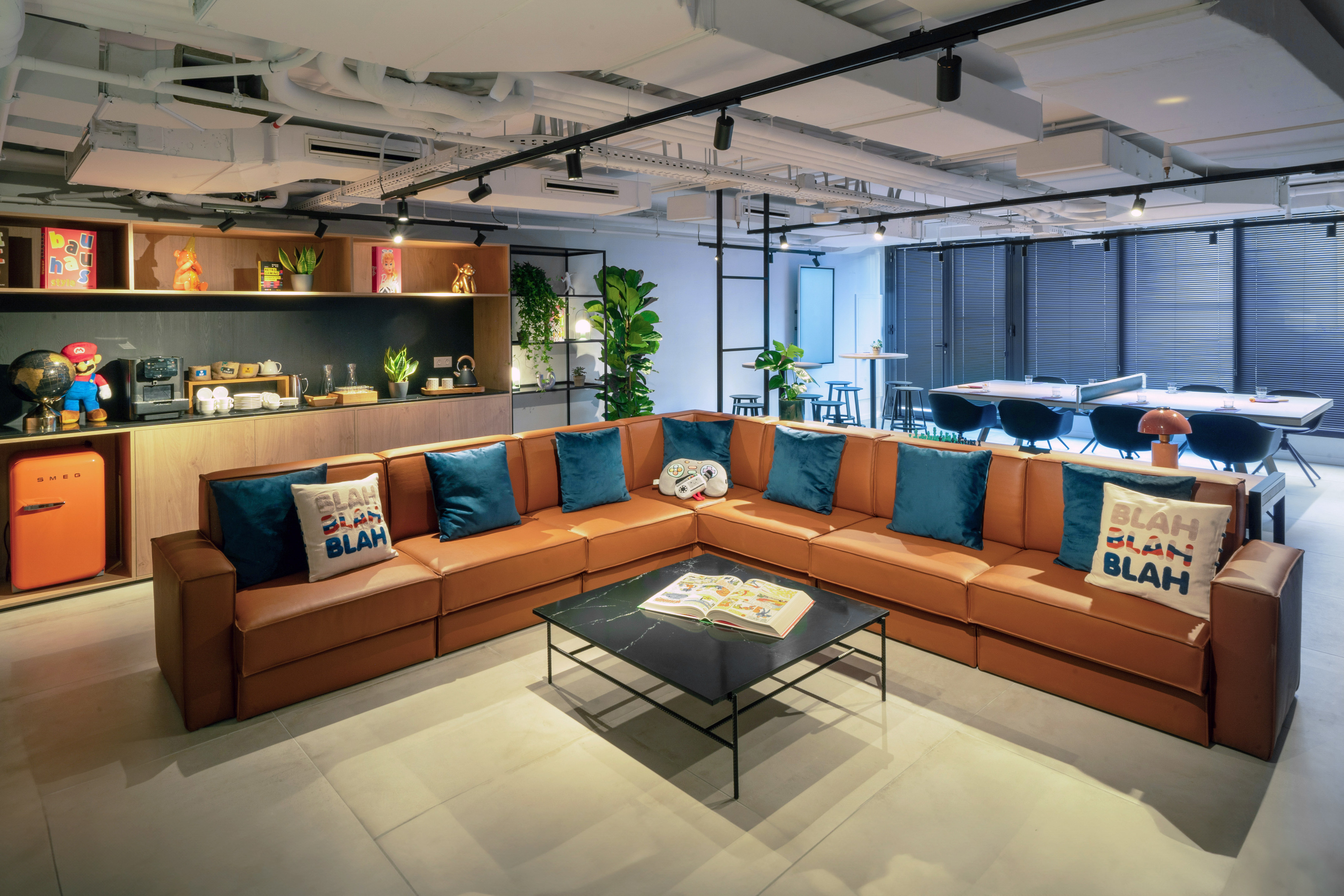 Modern office lounge with L-shaped brown sofa, kitchenette, and collaborative area.