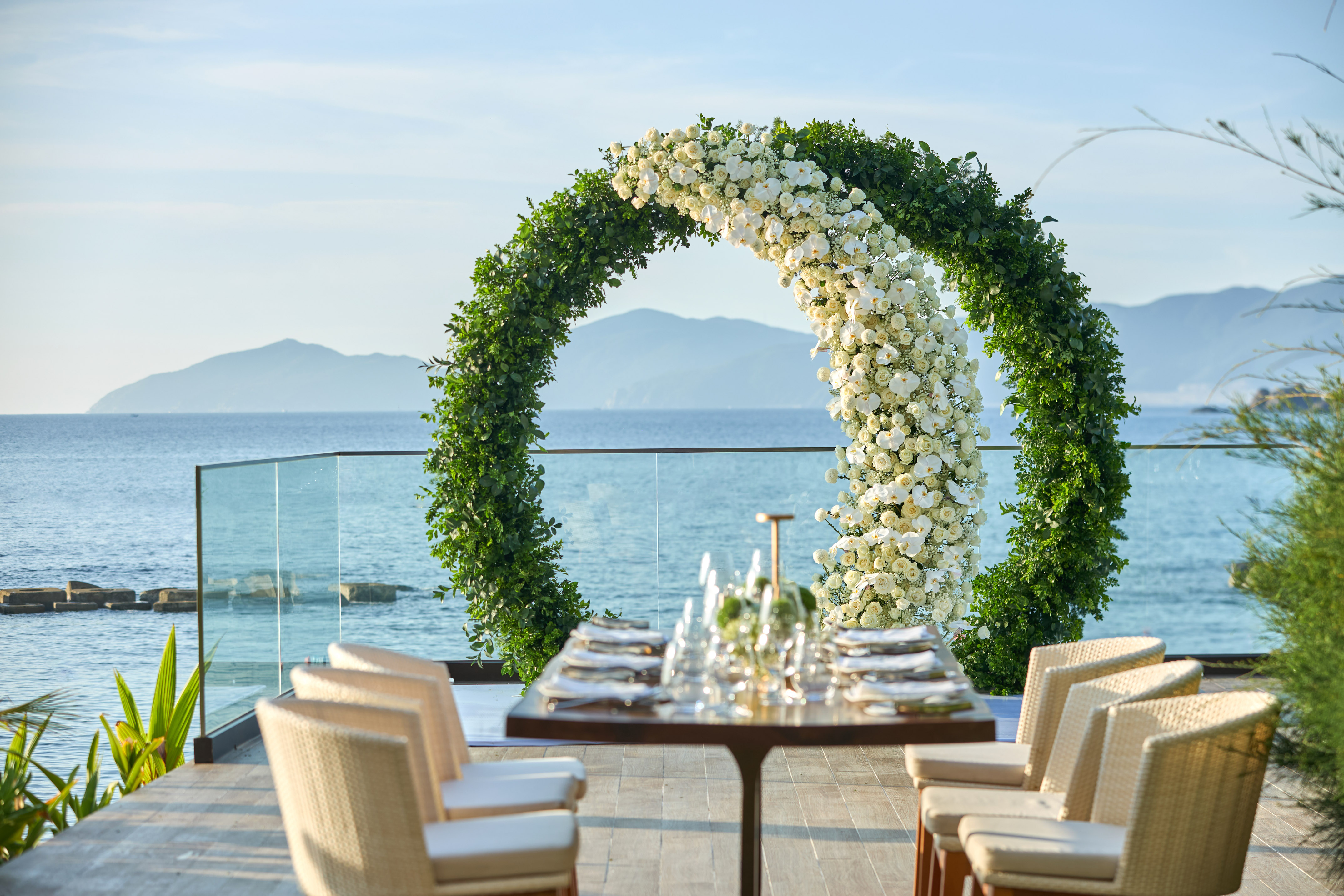 Oceanfront wedding arch, white floral & green, with elegant dining table.