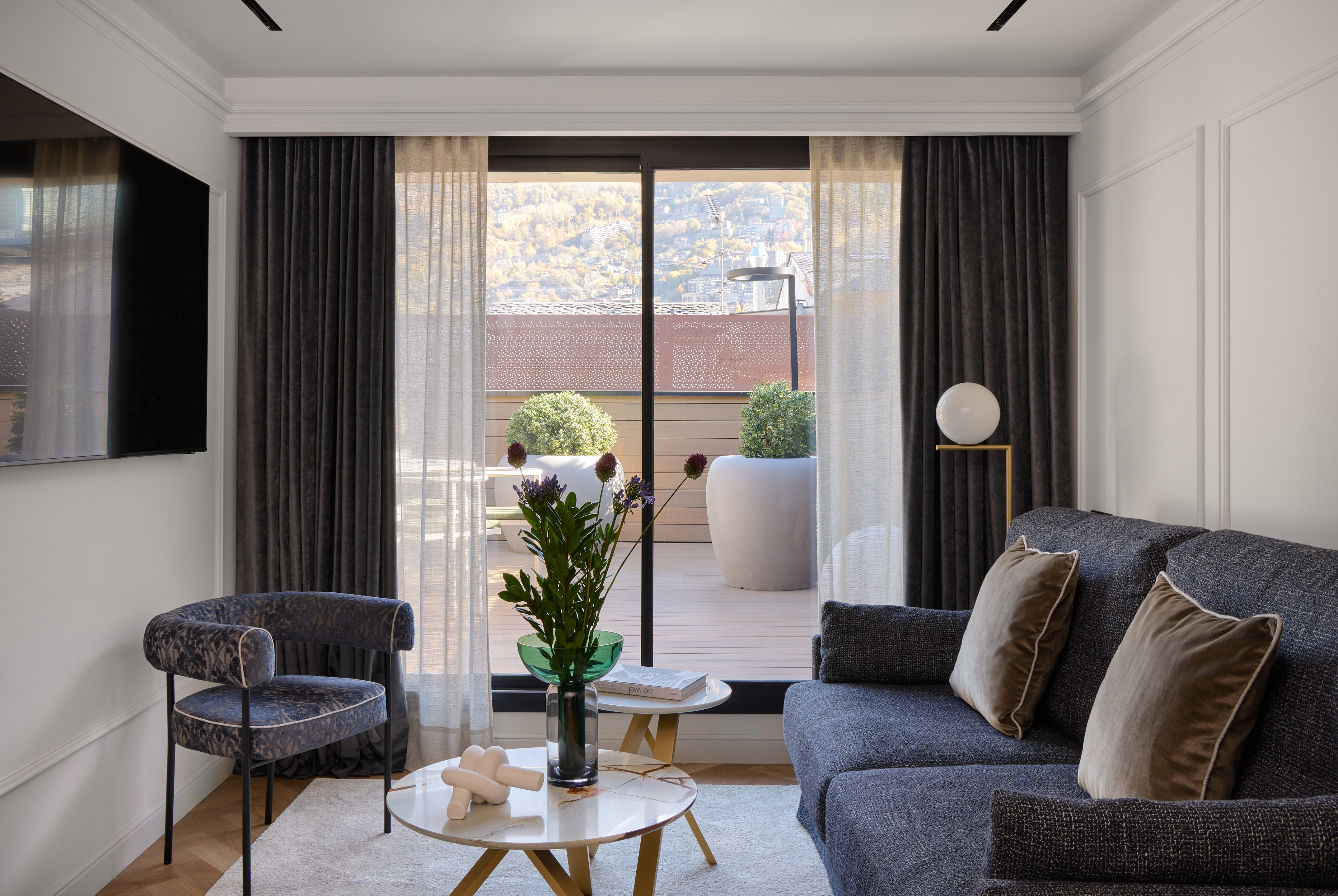 Modern living room with sofa, armchair, coffee tables, and city view from balcony.